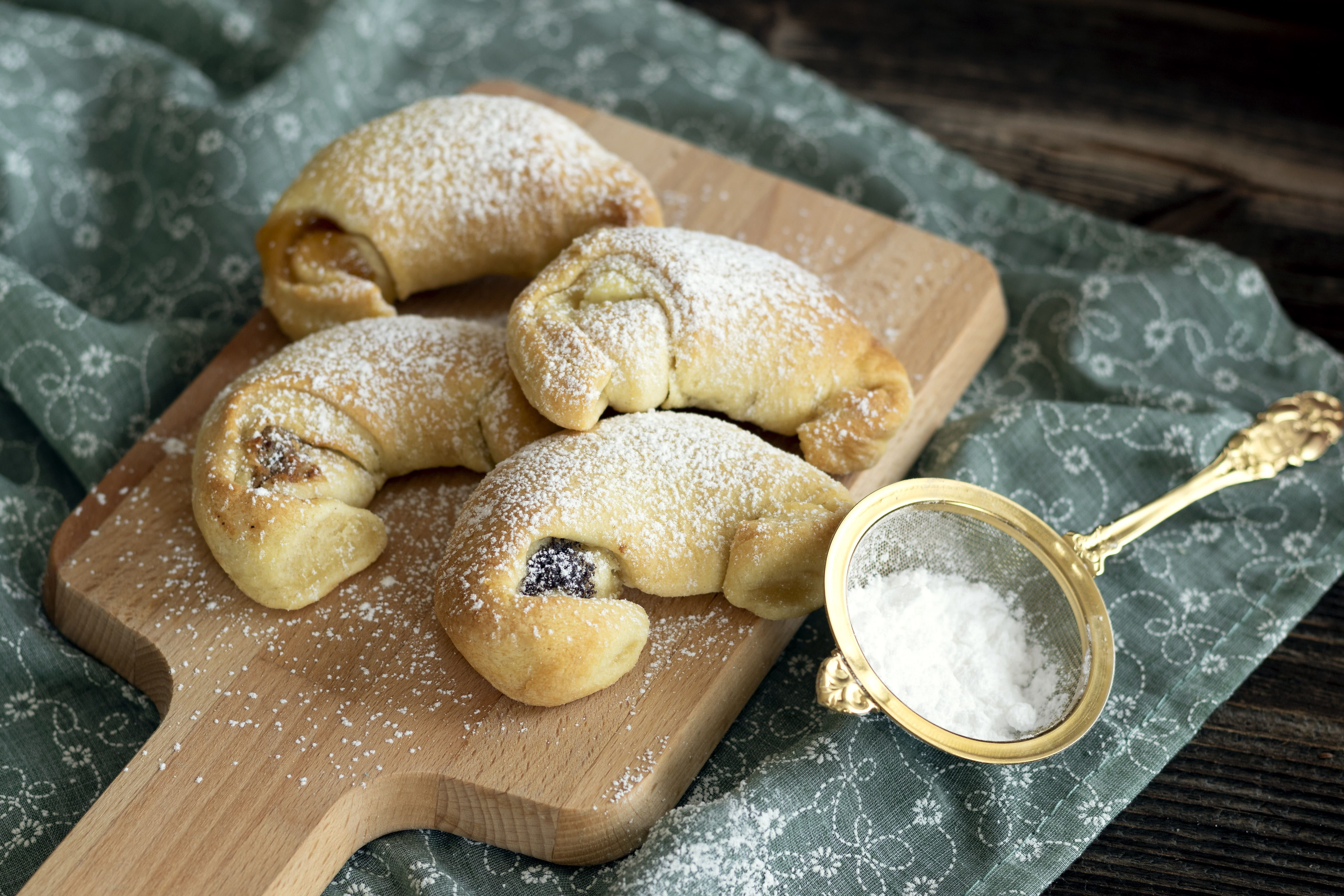 Čtyři krémové croissanty na dřevěné desce posypané moučkovým cukrem, vedle síta s moučkovým cukrem.