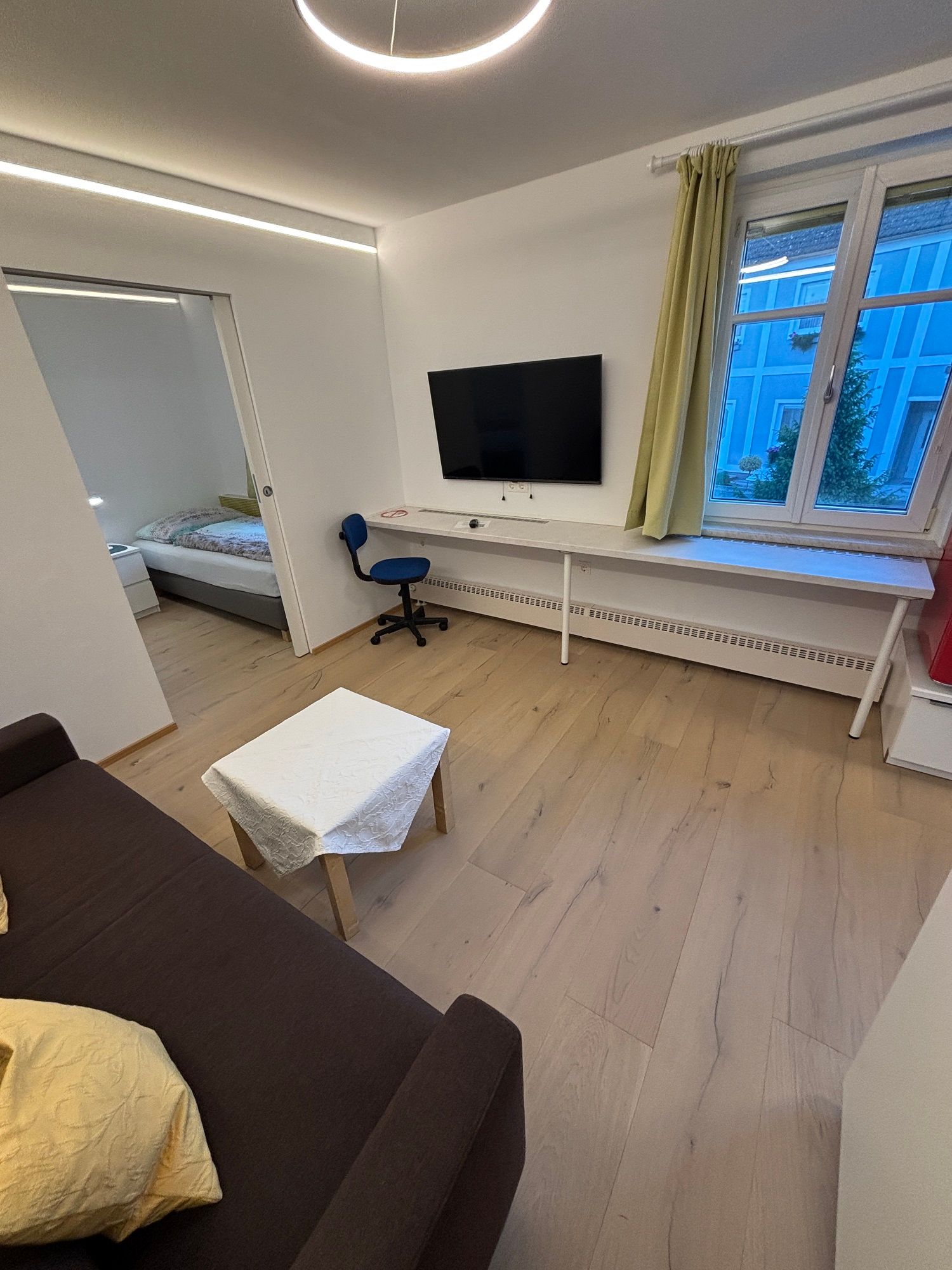 Moderní obývací pokoj s hnědou pohovkou, stolkem, televizí na stěně, psacím stolem a židlí. Okno s výhledem na modrou budovu.