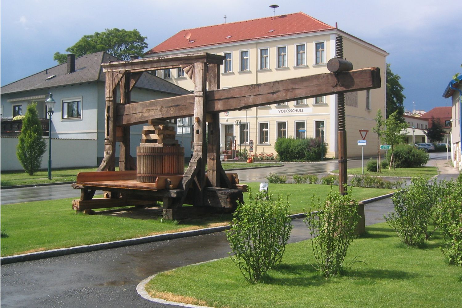 Historický lis na stromy před školní budovou v zeleném prostředí.