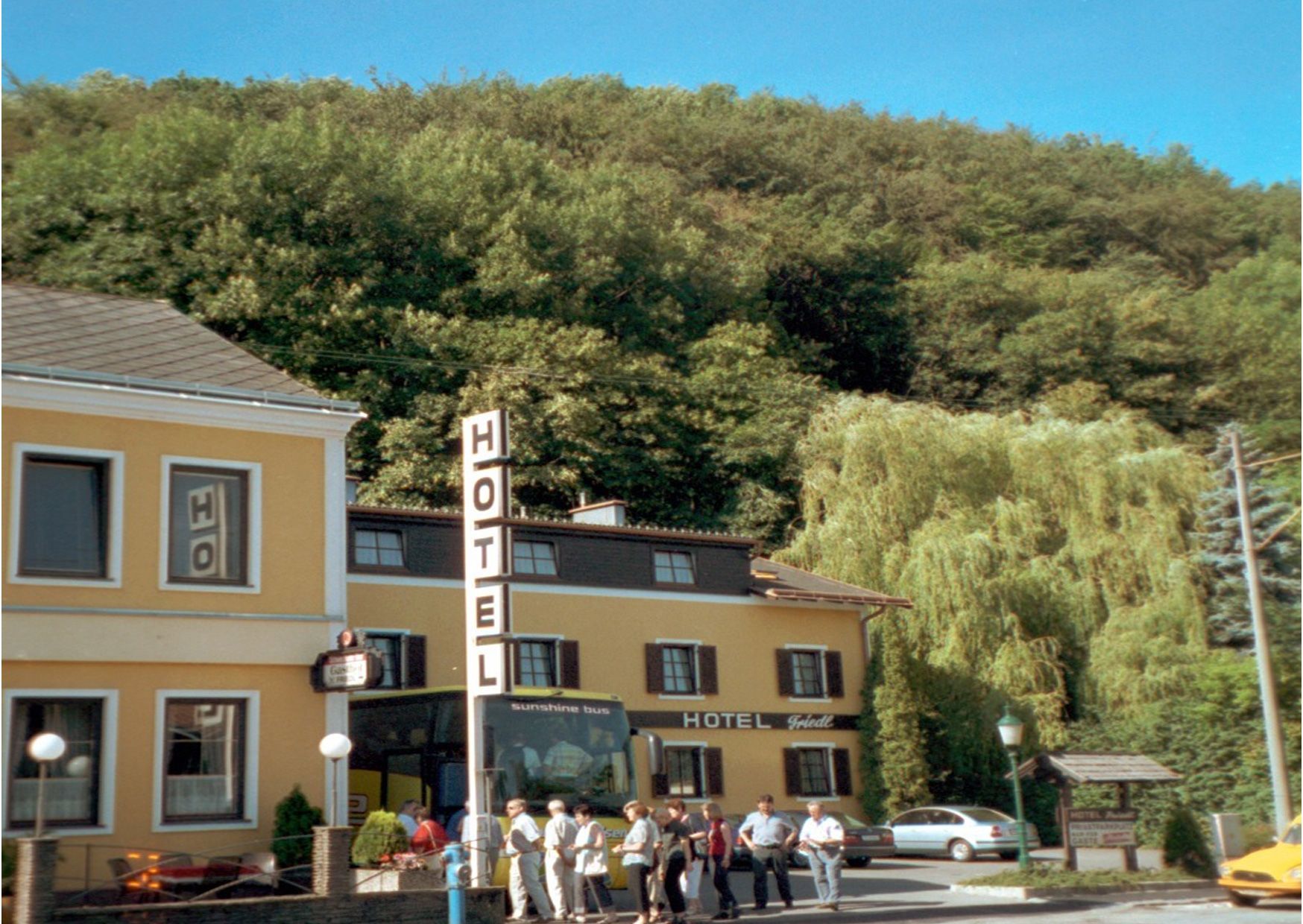 Žlutá budova hotelu s velkým nápisem a lidmi před ní, obklopená stromy.