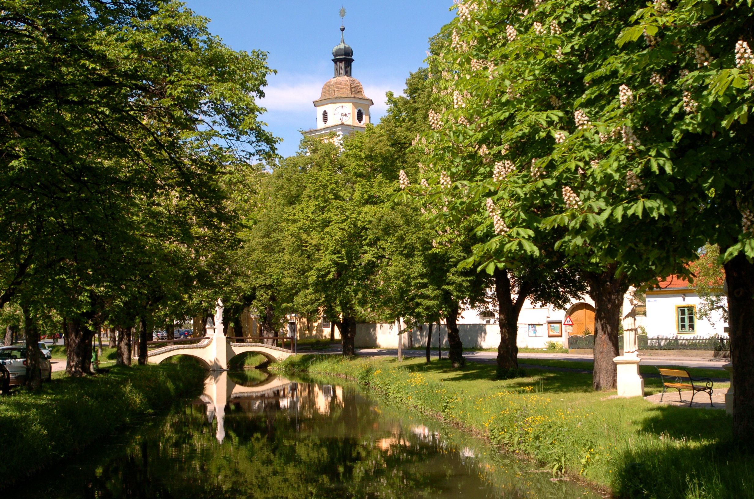 Idylický park s říčkou, mostem a kostelní věží v pozadí.