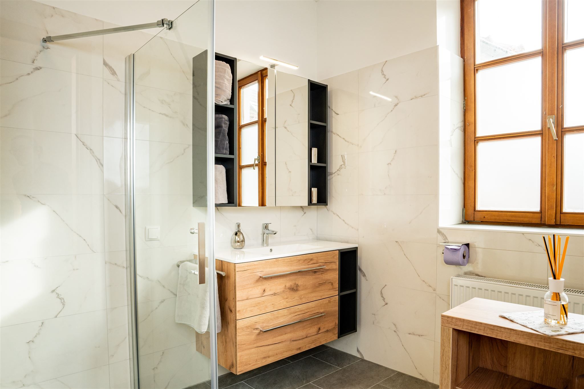 Moderní koupelna s dřevěným nábytkem, velkým čelním oknem a skleněným sprchovým koutem.