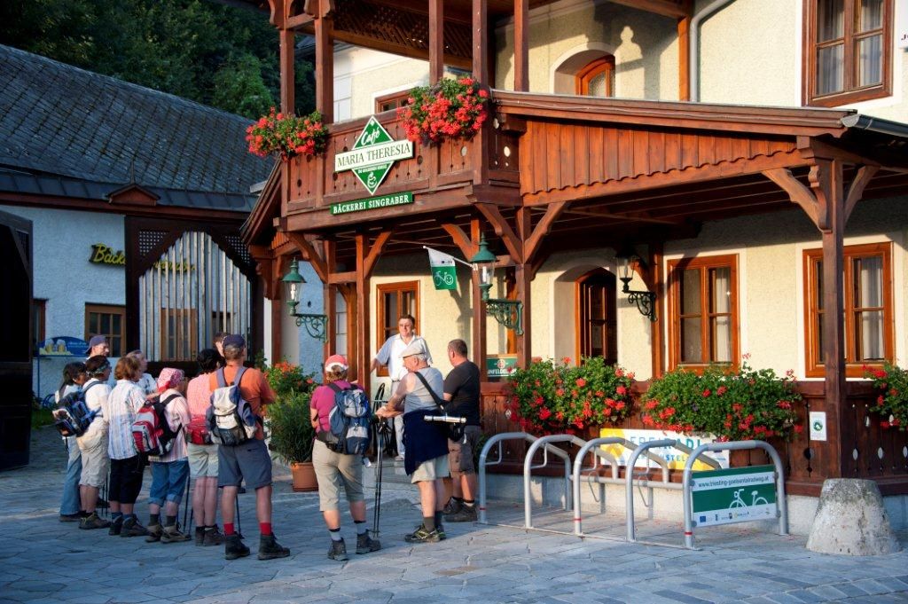 Skupina turistů stojí před kavárnou Maria Theresia s dřevěnou verandou a květinovou výzdobou.