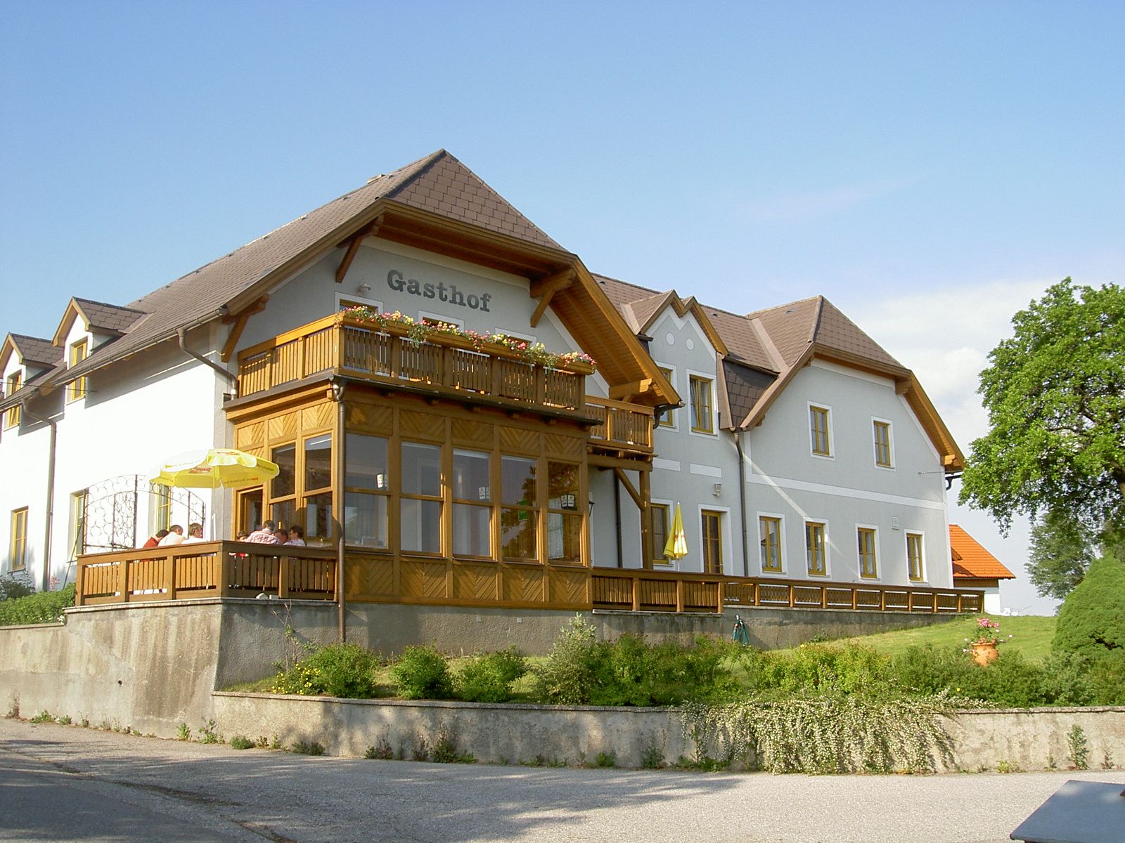 Tradiční hostinec s dřevěnou verandou a květinami na balkoně, obklopený zelenou krajinou.