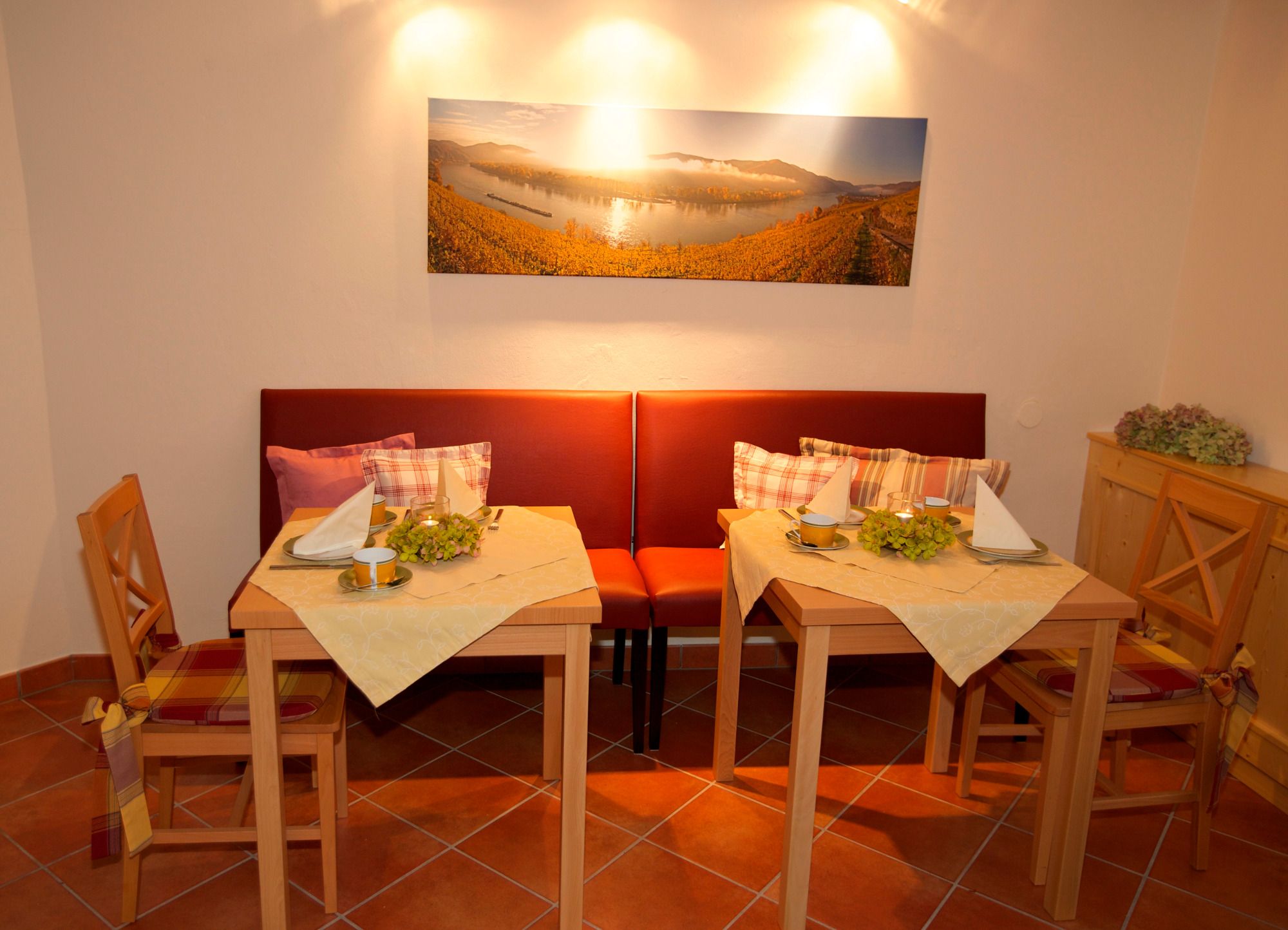 Útulná snídaňová místnost se dvěma prostřenými stoly, červenými lavicemi a obrazem krajiny na stěně.