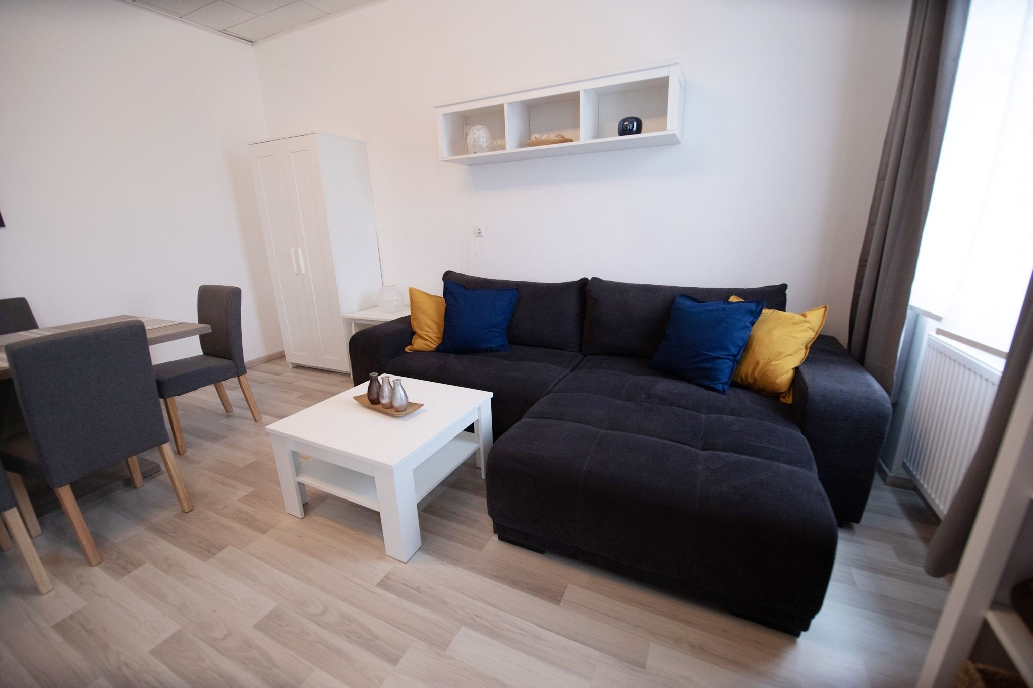 Moderní obývací prostor s šedou pohovkou, barevnými polštáři, bílým konferenčním stolkem a jídelním stolem se židlemi.