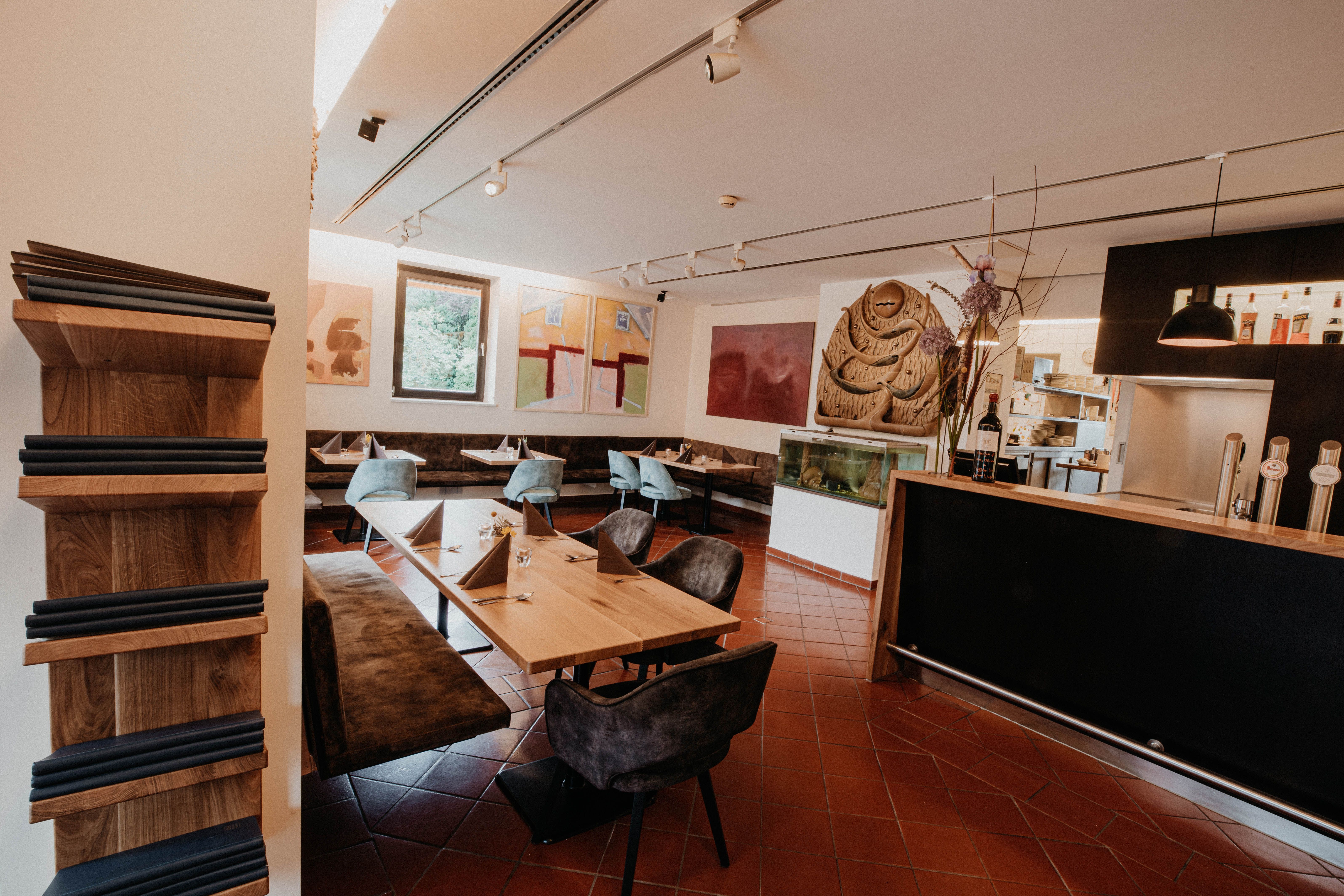 Pohled do interiéru moderní restaurace s dřevěnými stoly, modrými židlemi a uměleckými díly na stěnách.