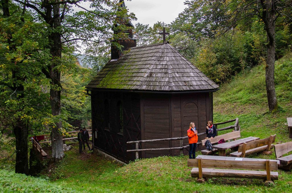 Malý dřevěný kostelík v lese se dvěma lidmi a dřevěnými lavicemi v popředí.