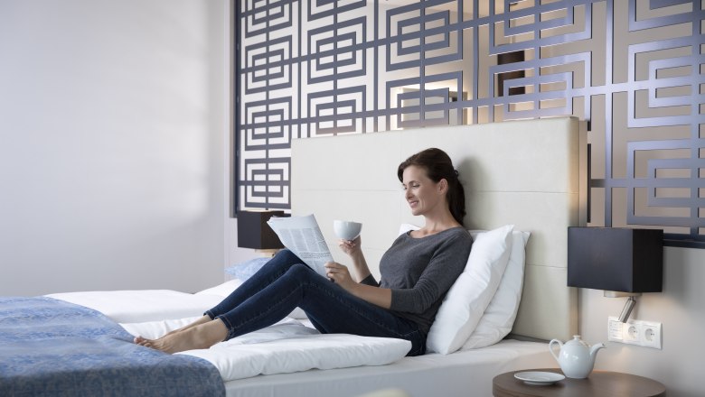 Žena odpoč&iacute;v&aacute; na posteli v modern&iacute;m hotelov&eacute;m pokoji, čte si noviny a drž&iacute; &scaron;&aacute;lek. Dekorativn&iacute; stěna v pozad&iacute;.