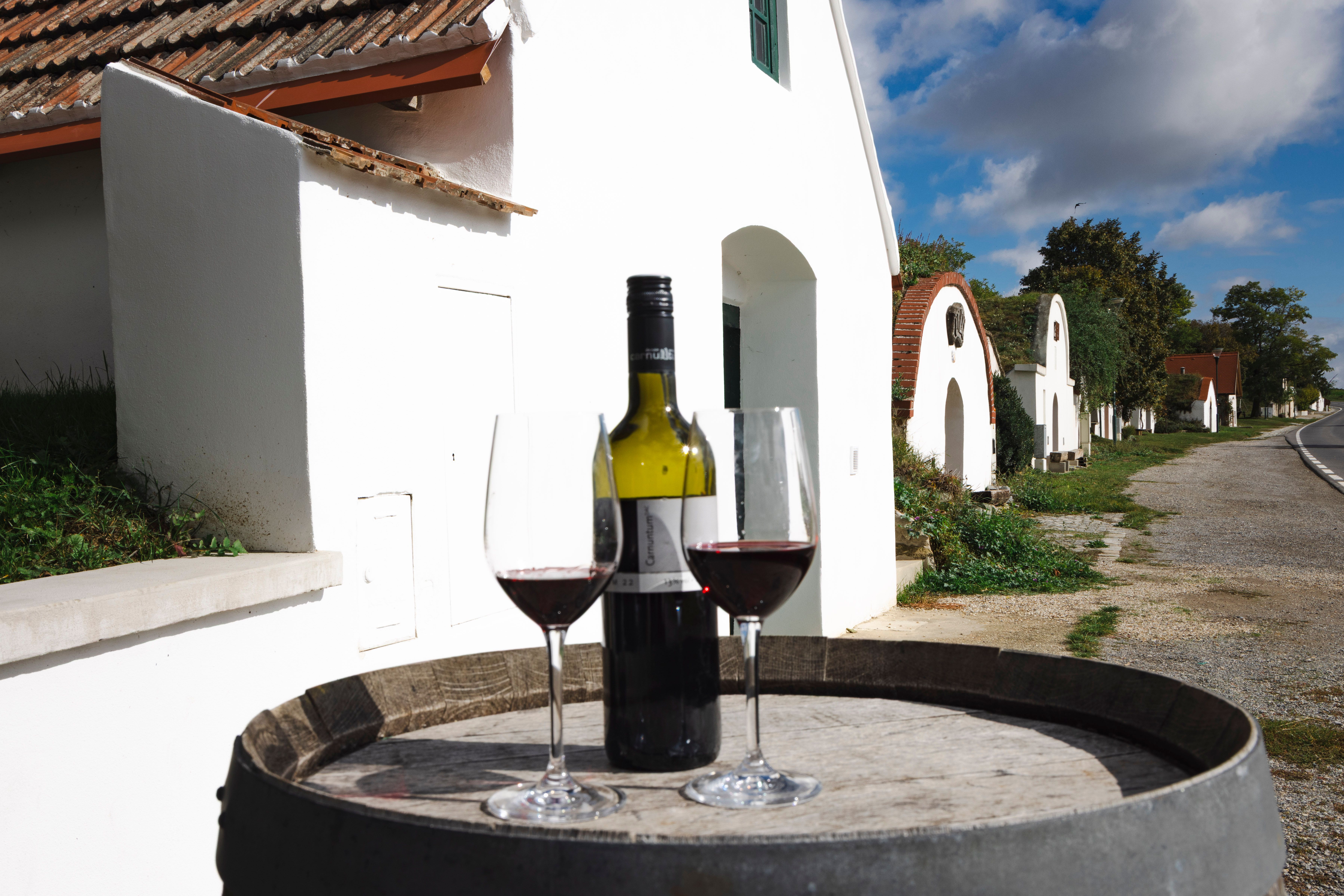 Dvě sklenice na víno a láhev červeného vína na sudu, v pozadí sklepní uličky