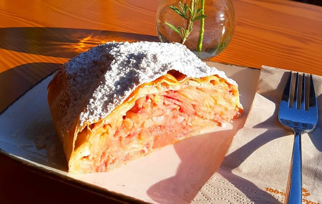 Na talíři kousek jablečného závinu s moučkovým cukrem, vedle vidlička a váza s rozmarýnem.