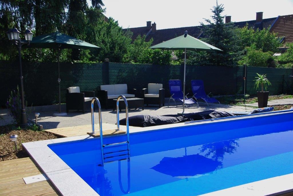Venkovní prostor s modrým bazénem obklopeným lehátky, slunečníky a zahradním nábytkem.