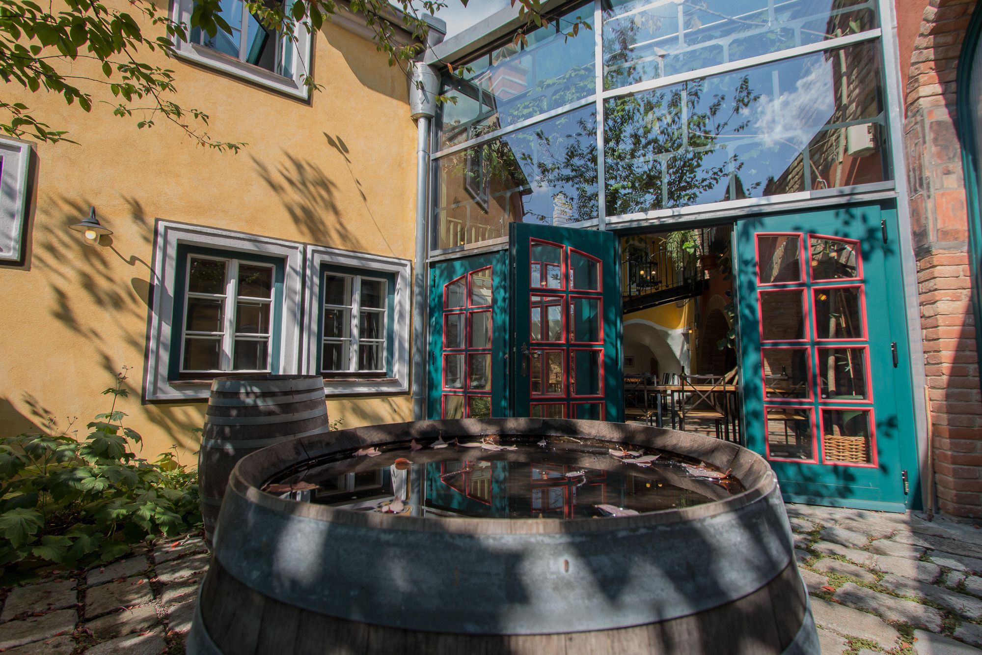 Vnitřní dvůr se žlutou fasádou, zelenými dveřmi a dřevěným sudem s vodou. Na stěnu dopadají stíny stromů.