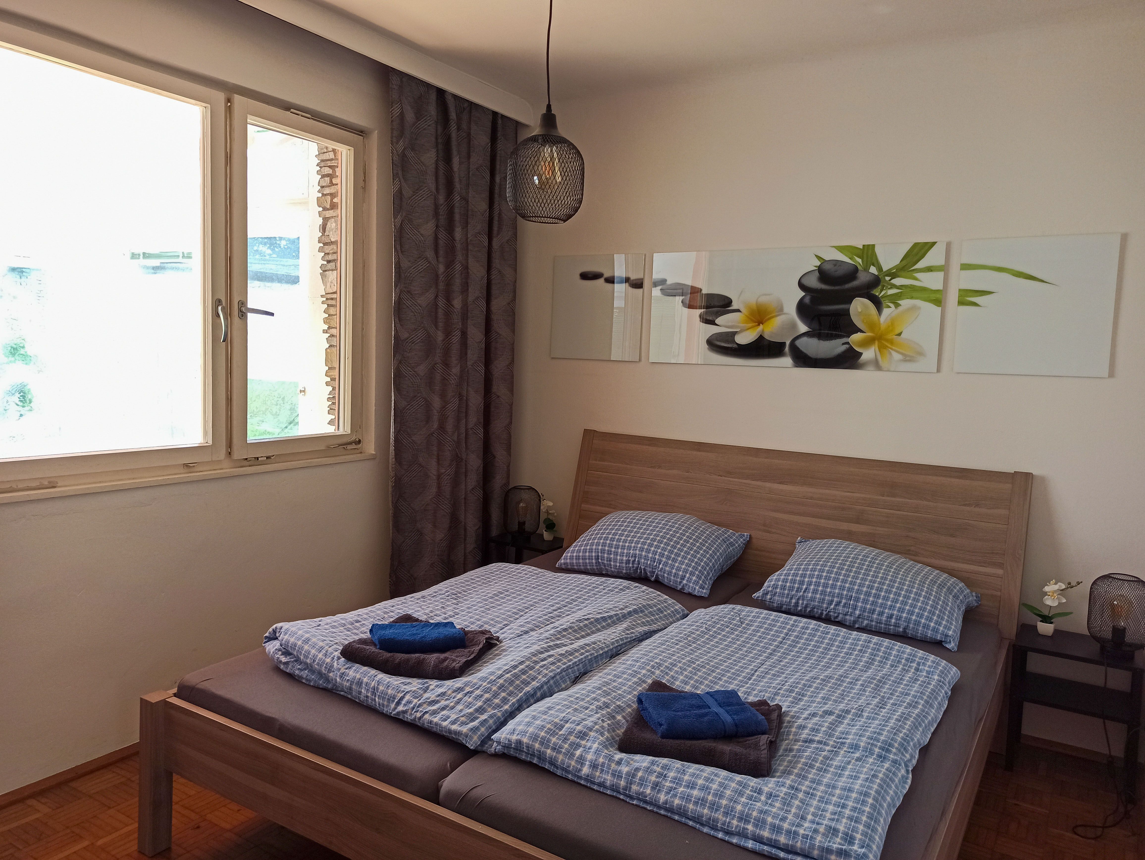 Ložnice s manželskou postelí, modré ložní prádlo, obraz s kameny a květinami na stěně, okno a noční lampička.