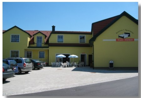 Žlut&aacute; budova s n&aacute;pisem "Pension Ziekel", před n&iacute; auta a stolky se slunečn&iacute;ky.