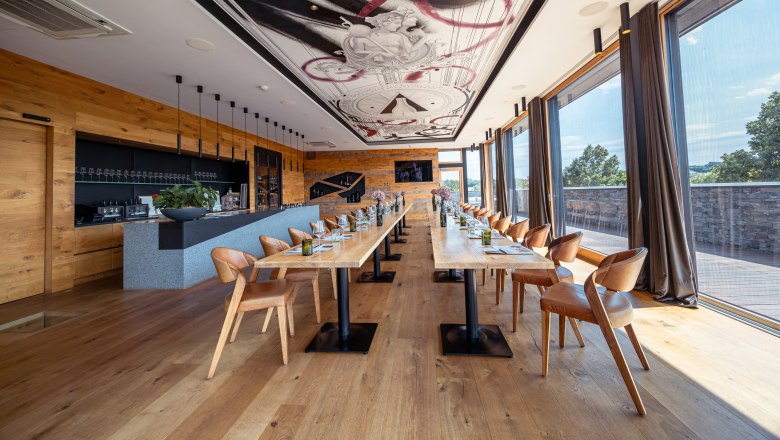 Pohled do interiéru moderní restaurace s dlouhými dřevěnými stoly, koženými křesly a velkými okny.