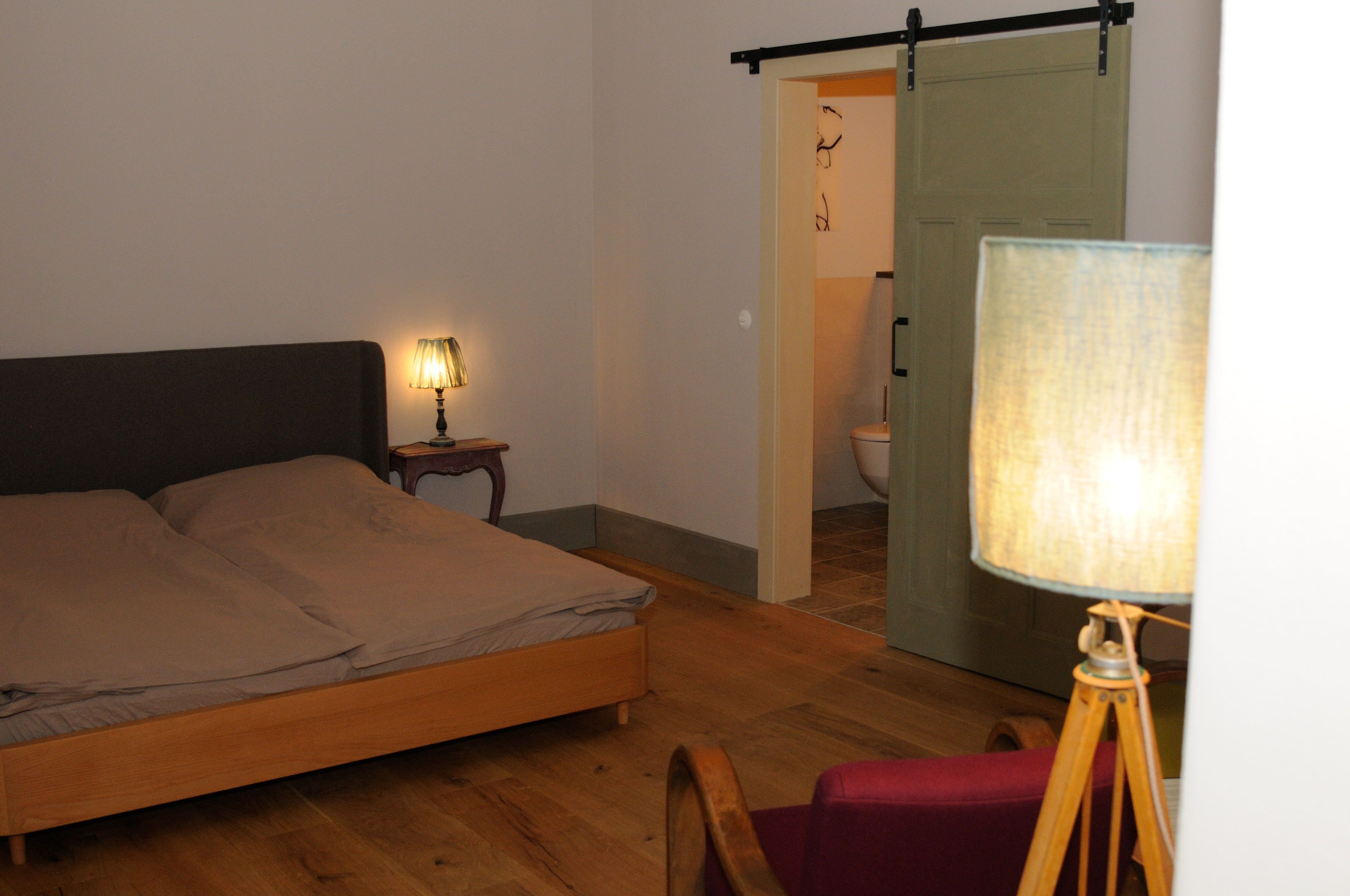 Útulná ložnice s manželskou postelí, noční lampičkou a posuvnými dveřmi do koupelny.