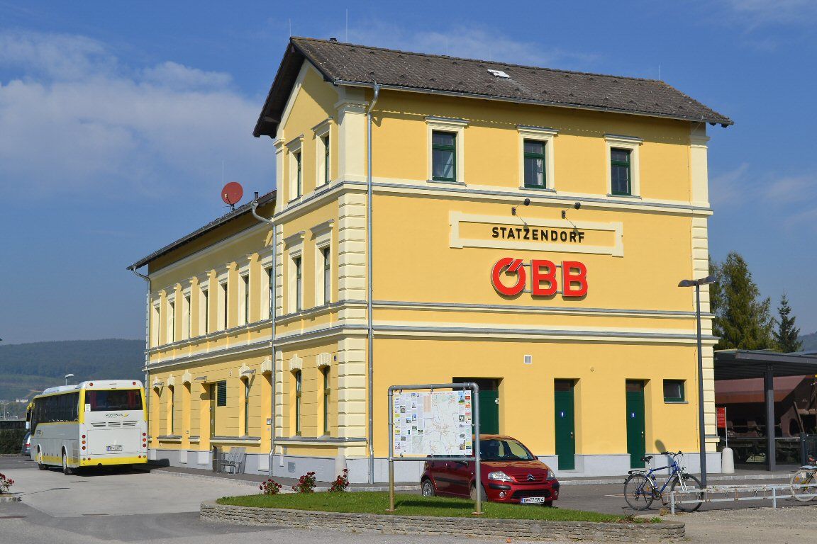 Žlutá nádražní budova s logem ÖBB a autobusem v popředí.