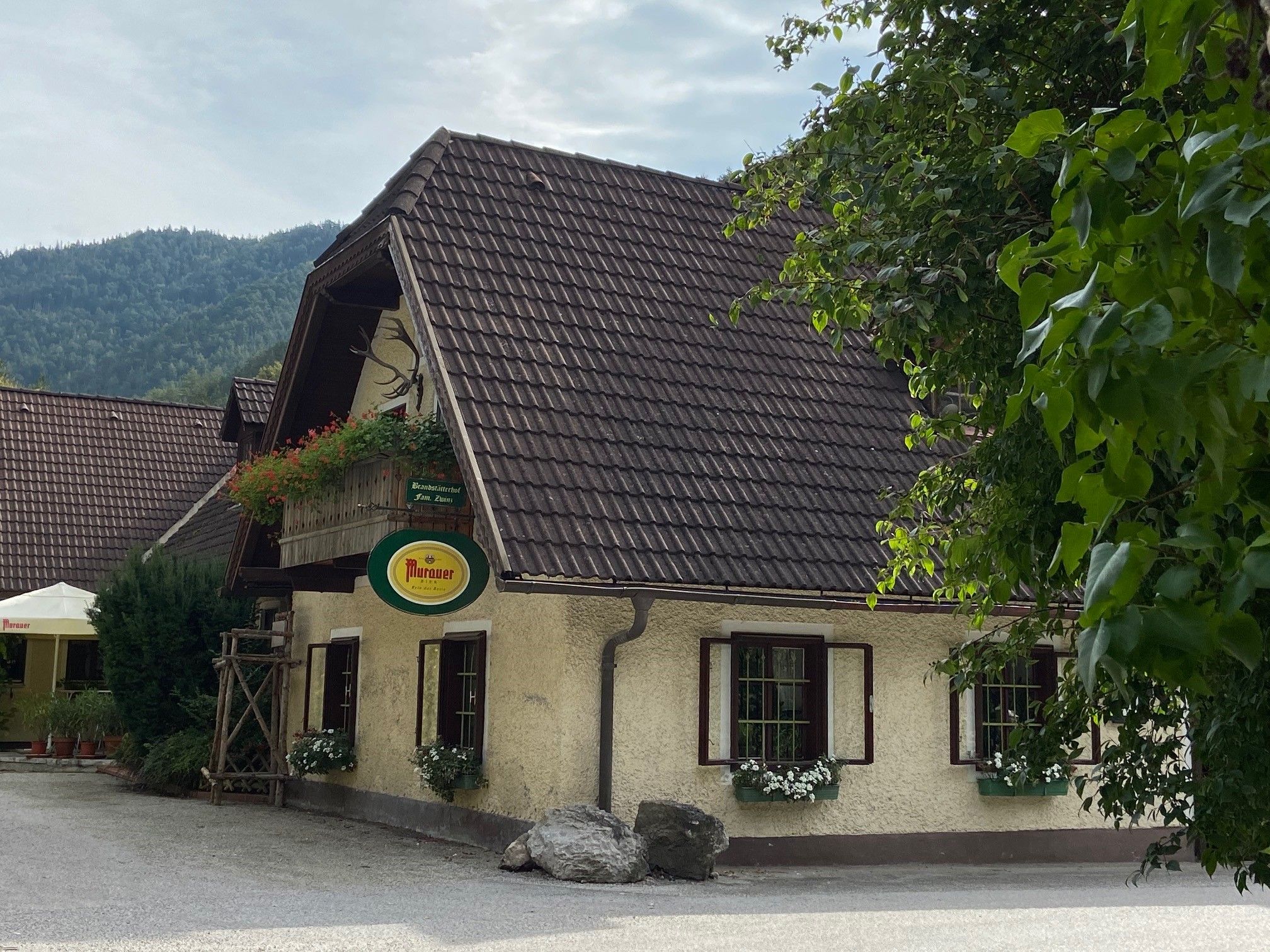 Tradiční hostinec s květinami a nápisem Murauer, obklopený stromy.