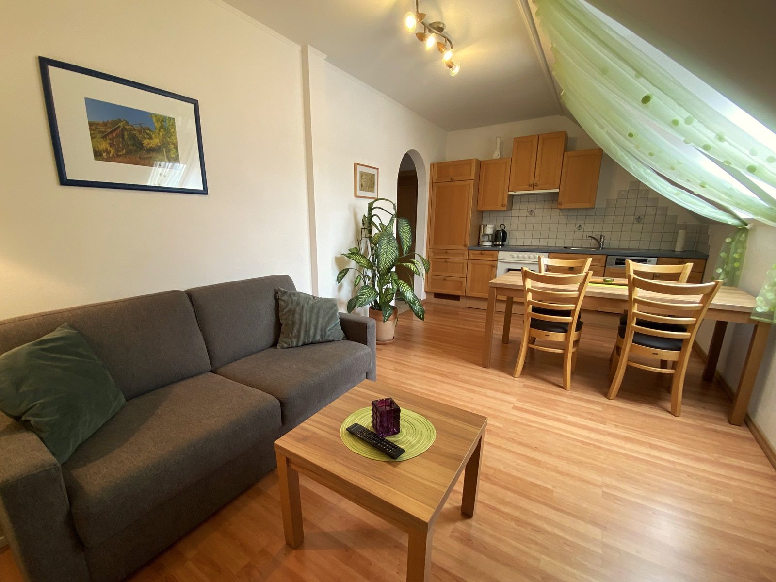 Obývací pokoj s šedou pohovkou, dřevěným stolem, jídelním stolem a kuchyní v pozadí.