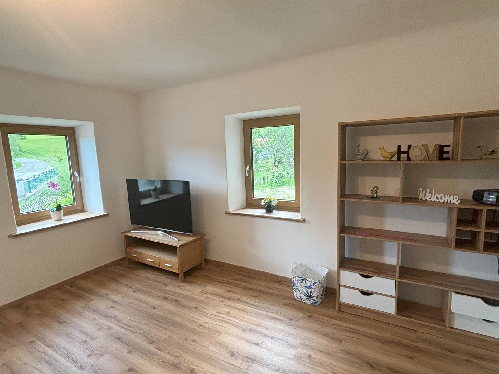 Obývací pokoj s dřevěnou podlahou, televizí na stole, dvěma okny a policí s dekoracemi.