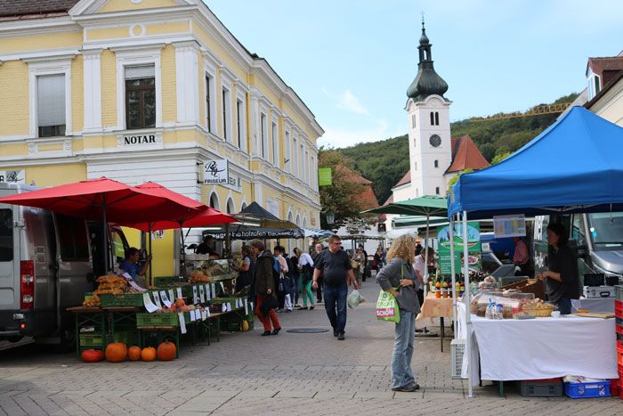 Živý farmářský trh se stánky a návštěvníky před žlutou budovou a kostelem v pozadí.