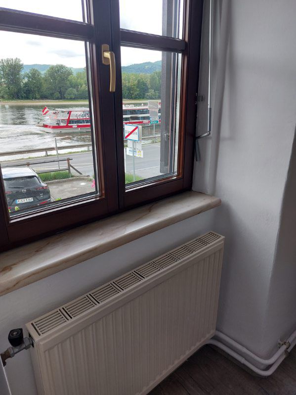 Pohled z okna na řeku s projíždějící lodí a zaparkovaným autem.