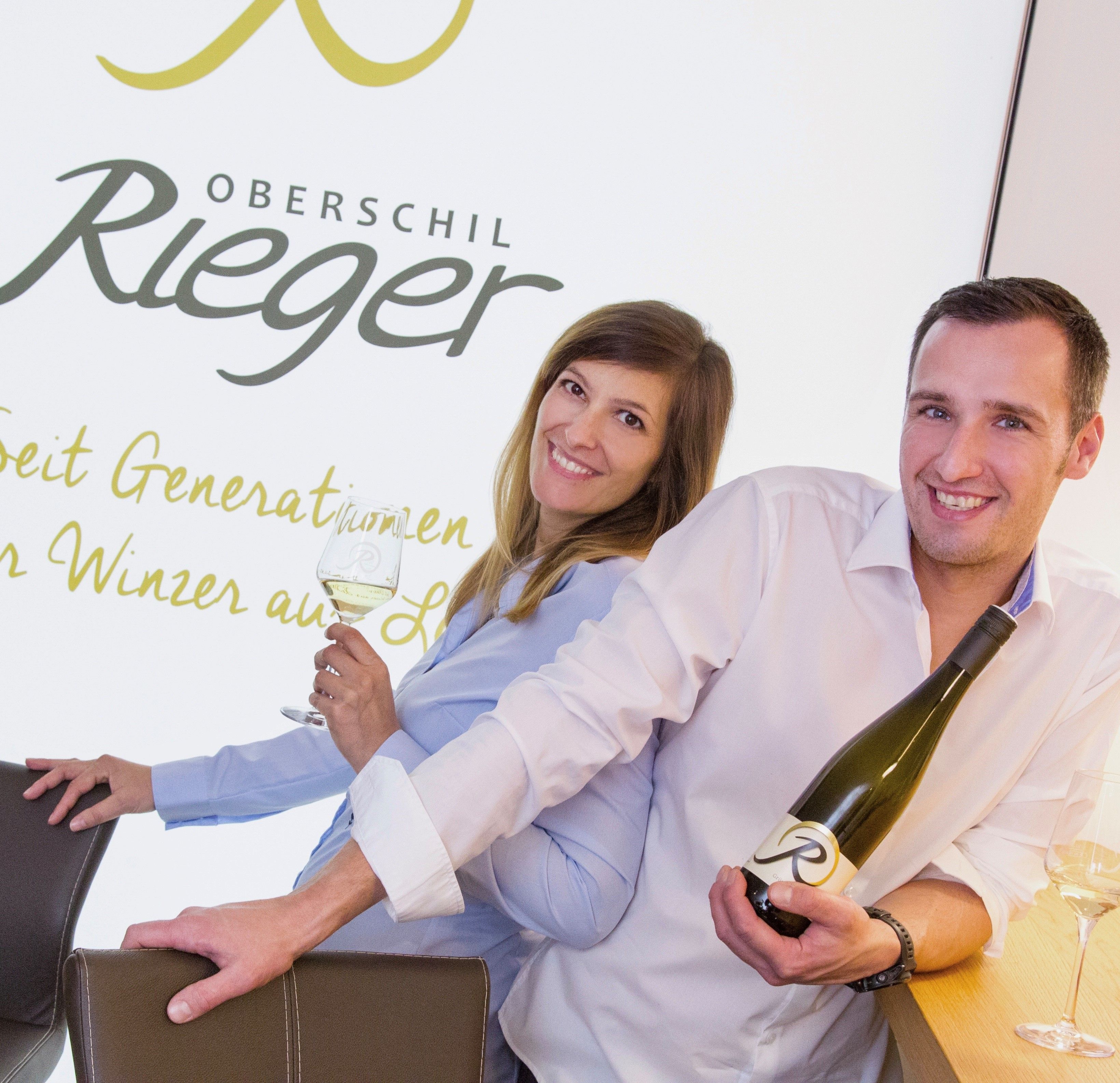 Dva lidé se usmívají a drží láhve vína před nápisem "Oberschil Rieger".