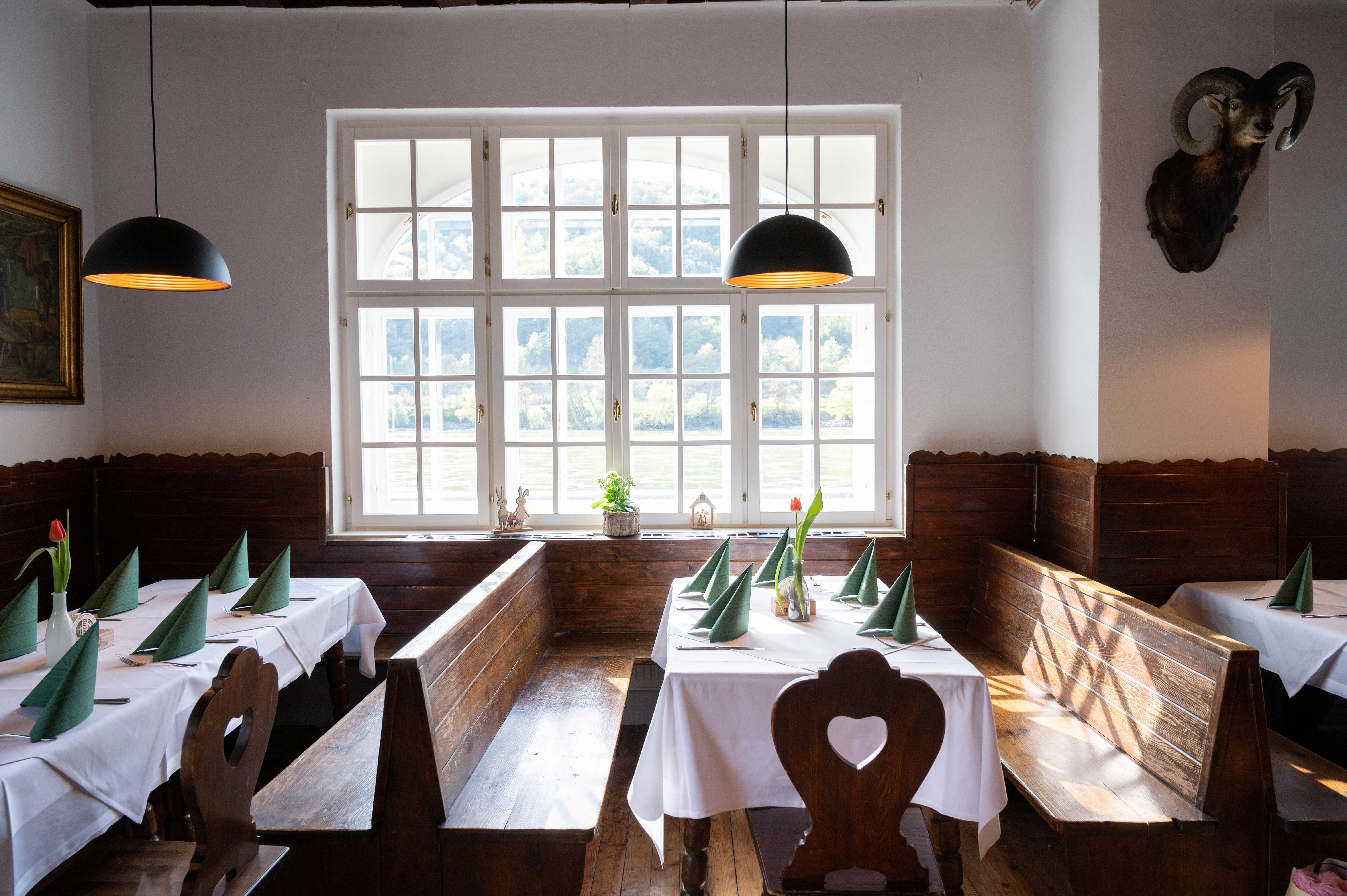 Útulná jídelna s dřevěnými lavicemi, bílými ubrusy a zelenými ubrousky. Velká okna s výhledem do přírody.