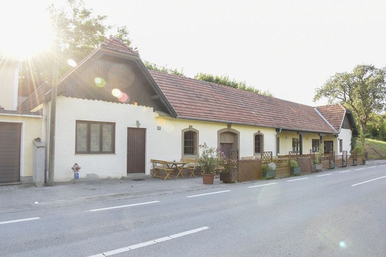 Tradiční budova s červenou taškovou střechou a dřevěnými stoly před vchodem, osvětlená sluncem.