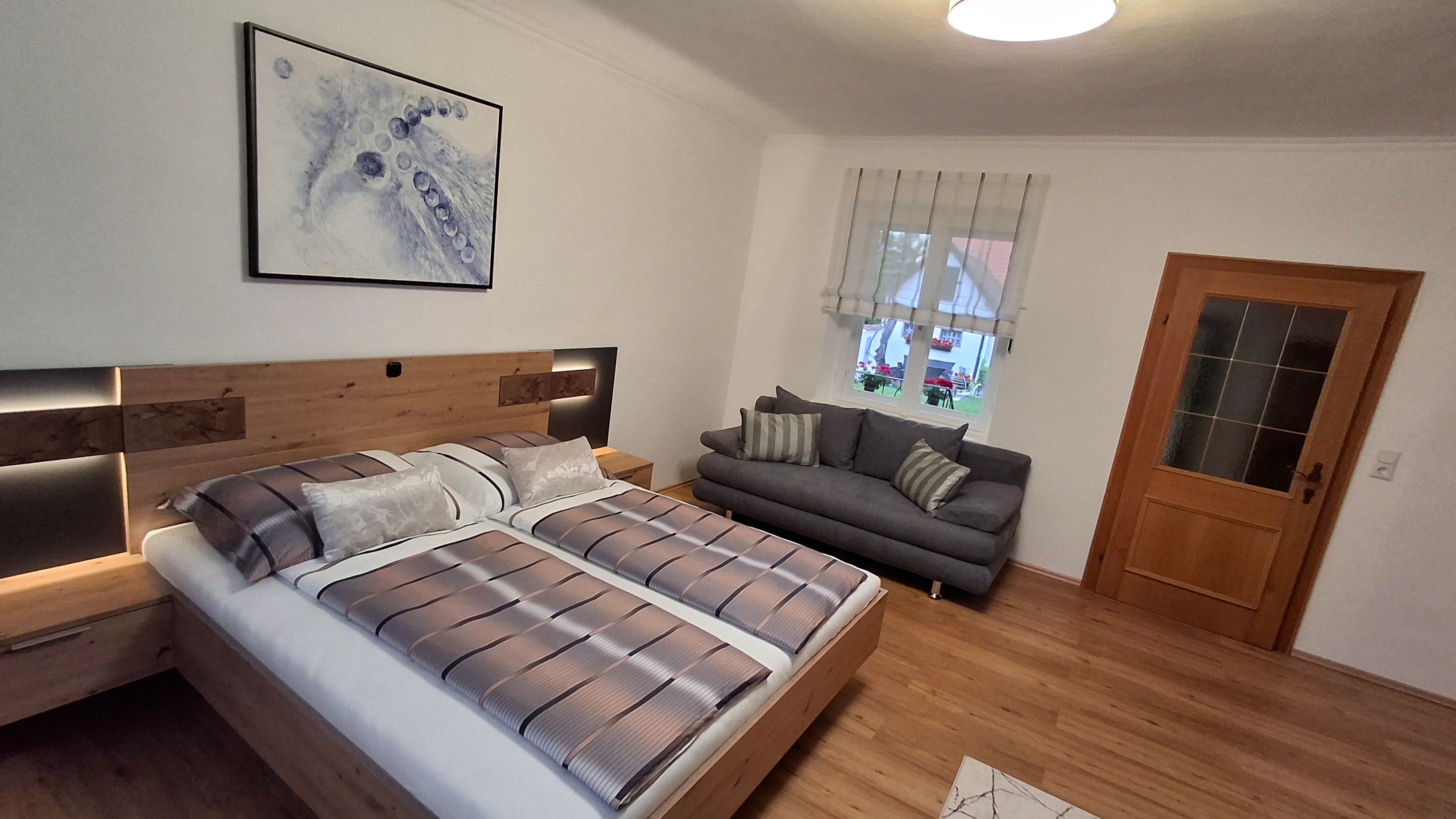 Moderní ložnice s manželskou postelí, šedou pohovkou, dřevěnou podlahou a abstraktním uměleckým dílem na stěně.