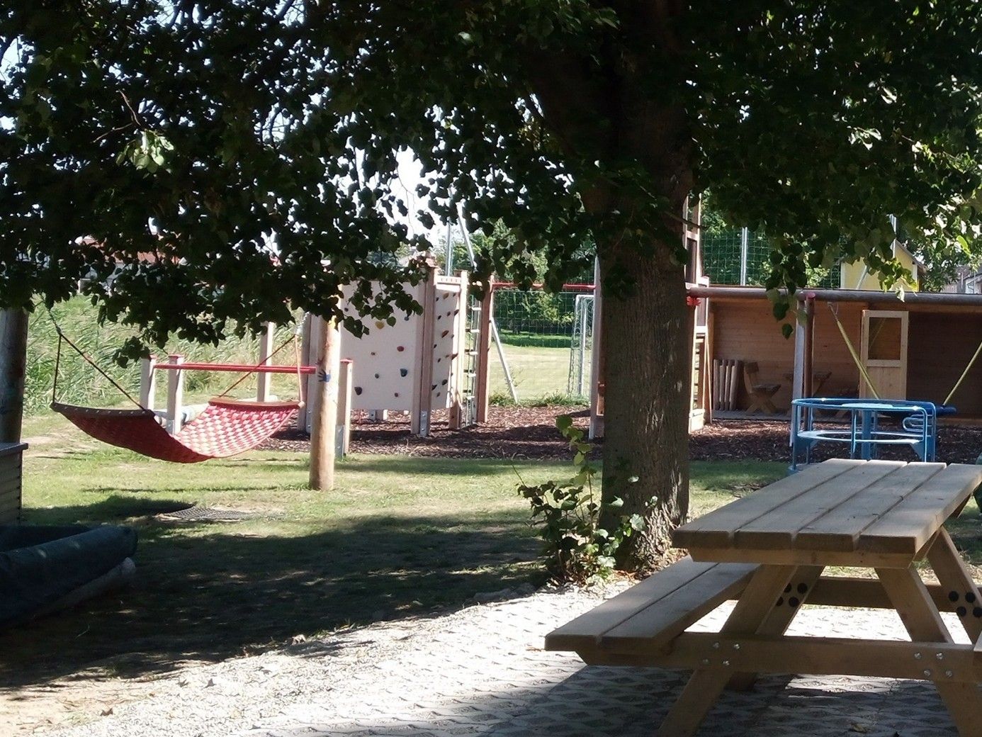 Dětské hřiště s houpací sítí, lezeckou stěnou a dřevěným stolem pod stromem.