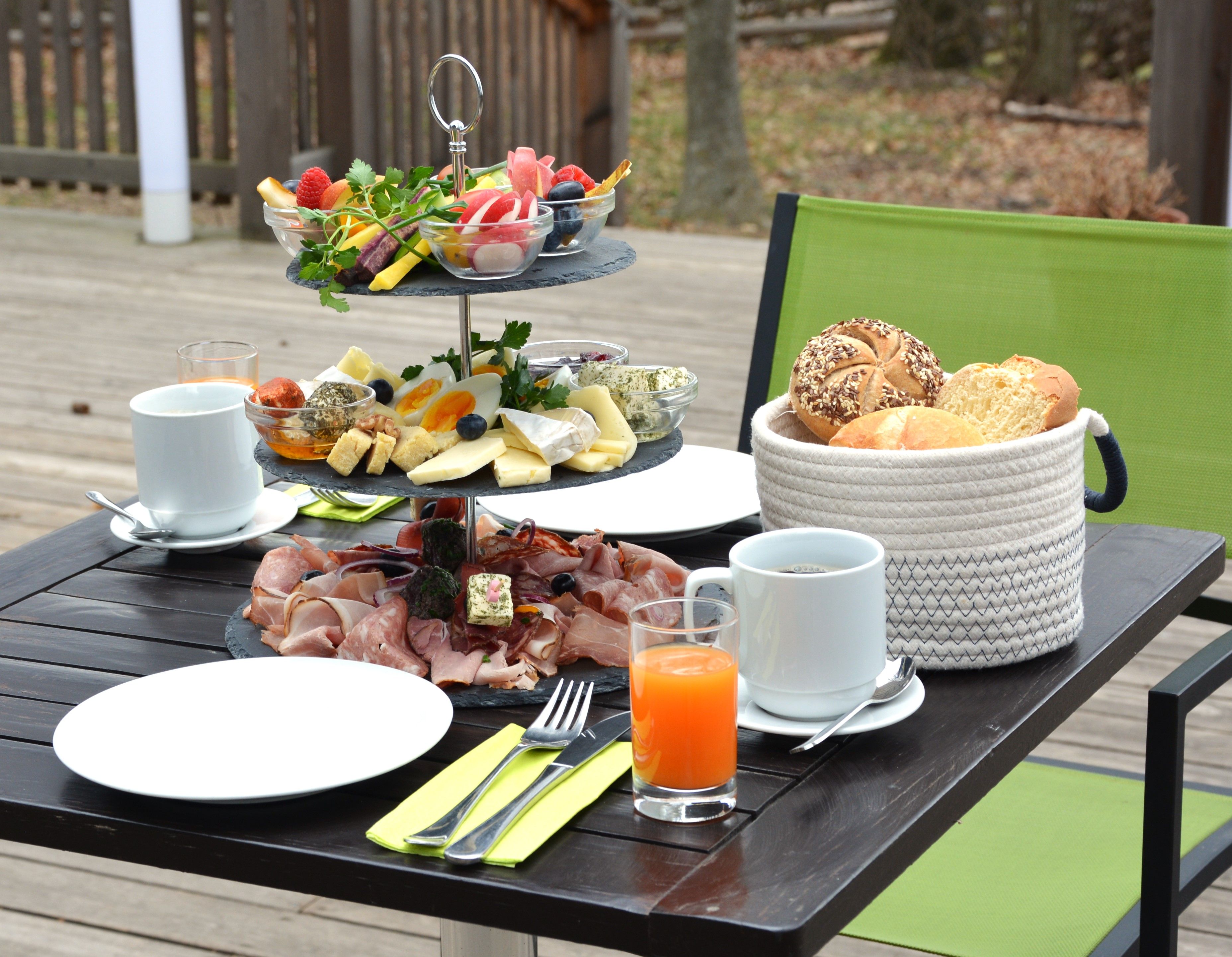 Snídaňový stůl venku s uzeninami, sýrem a zeleninou. Vedle něj talíře, příbory, nápoje a košík na pečivo.