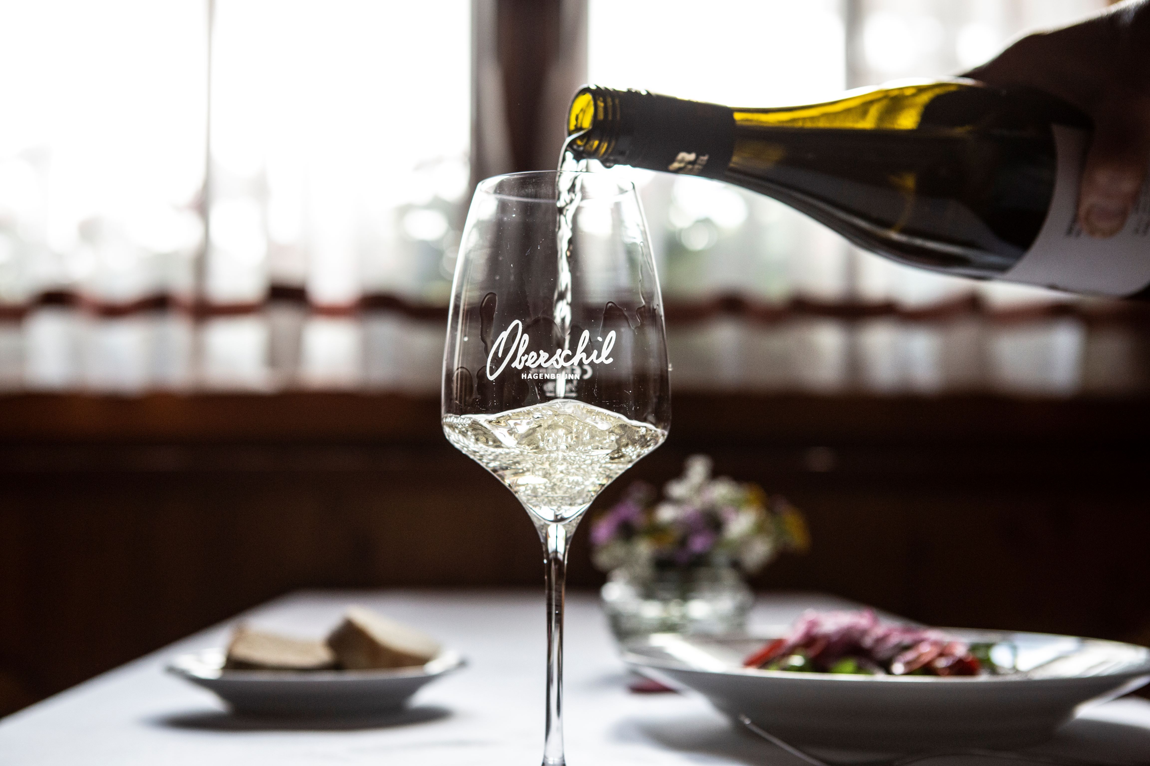 Nalévá se sklenka bílého vína, v pozadí je prostřený stůl s květinami.