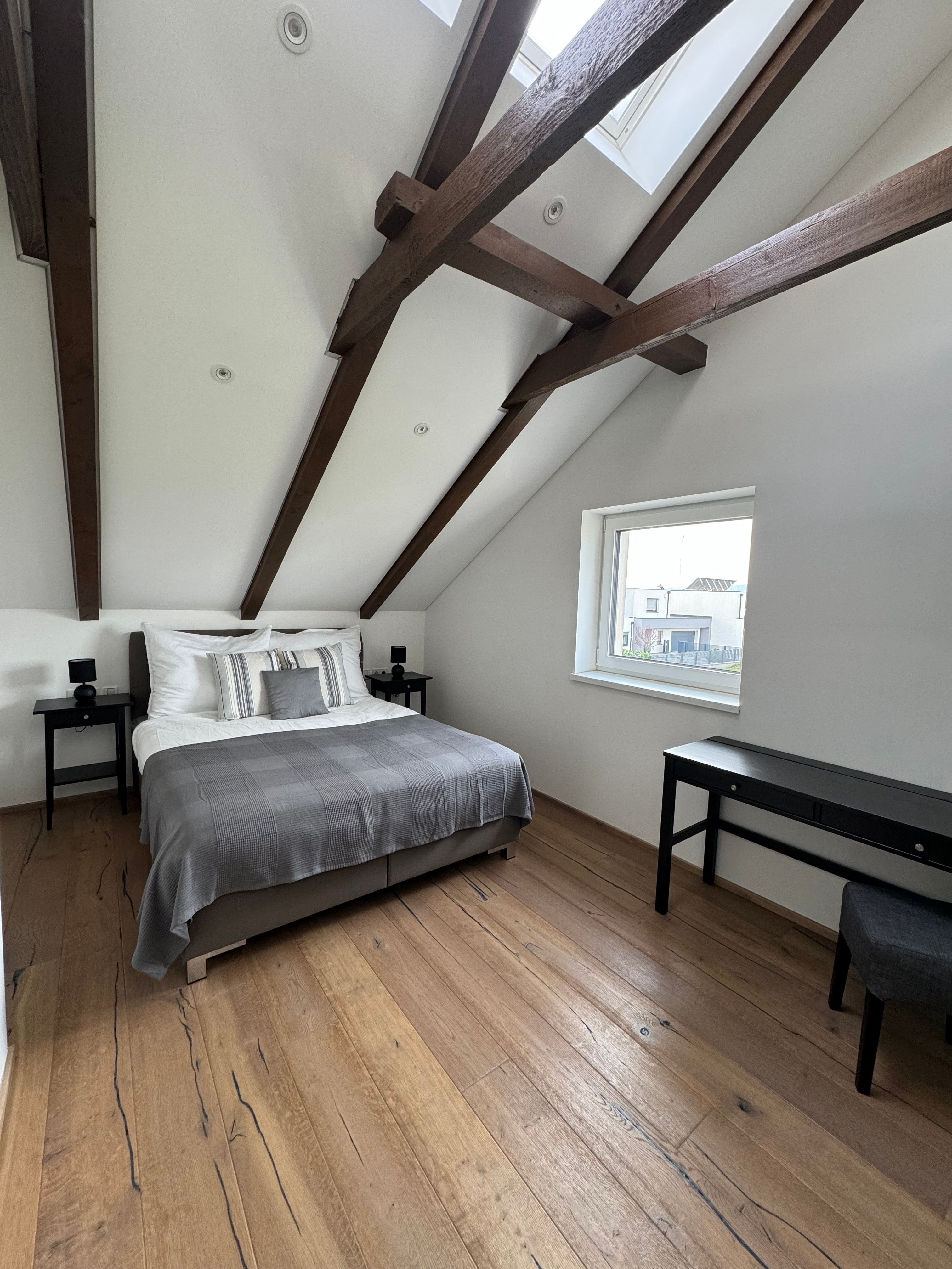 Moderní ložnice s dřevěným trámovým stropem, manželskou postelí, dřevěnou podlahou a oknem.