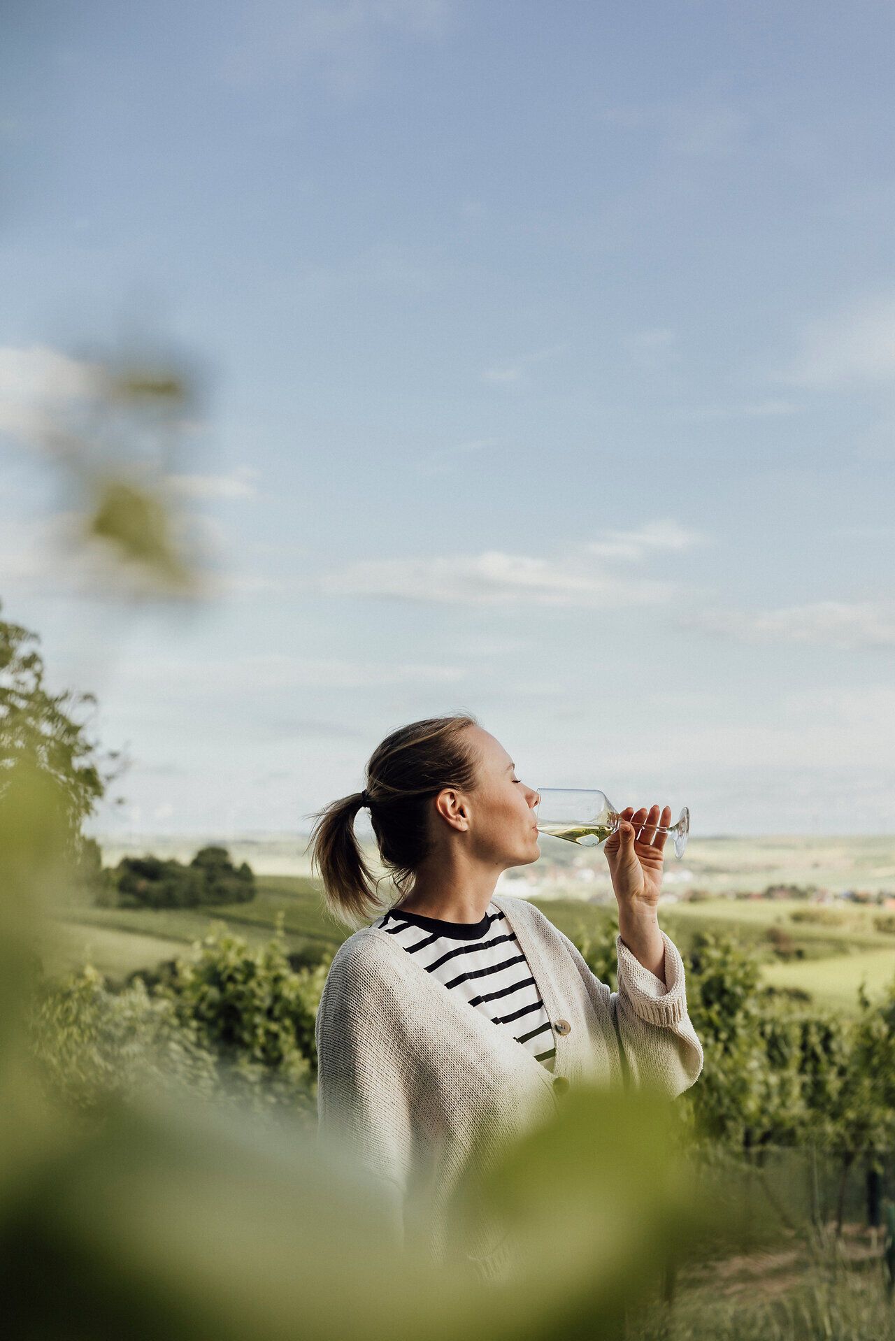 Inmitten der sanften Hügel des Weinviertels genießt eine Frau ein Glas Wein, während die grüne Landschaft sich weitläufig erstreckt. Der klare Himmel und die frische Luft laden dazu ein, den Moment der genussvollen Gelassenheit zu erleben und die Schönheit des Weinfrühlings zu feiern.