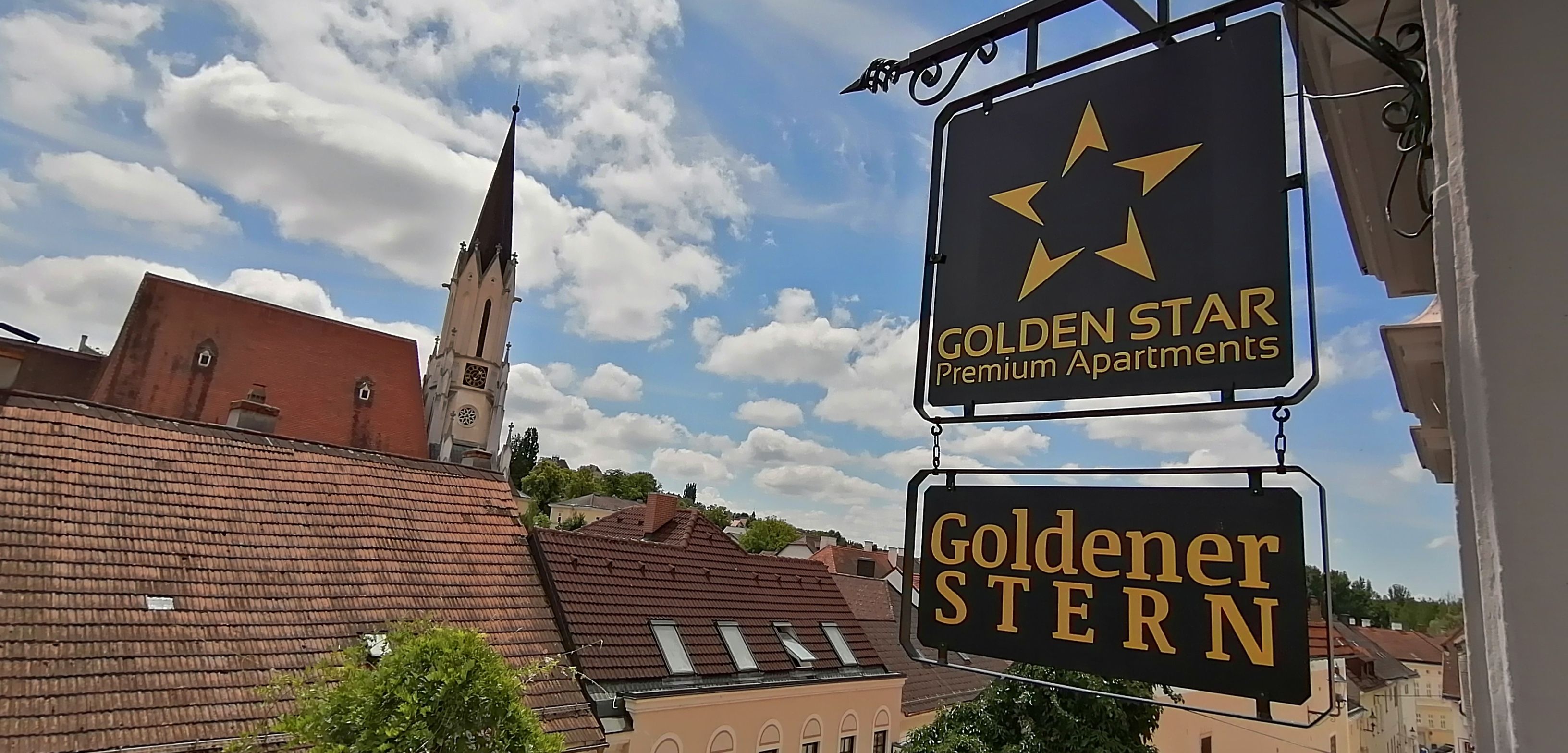 Pohled na ceduli s nápisem "Golden Star Premium Apartments" a "Goldener Stern" před kostelní věží a střechami.