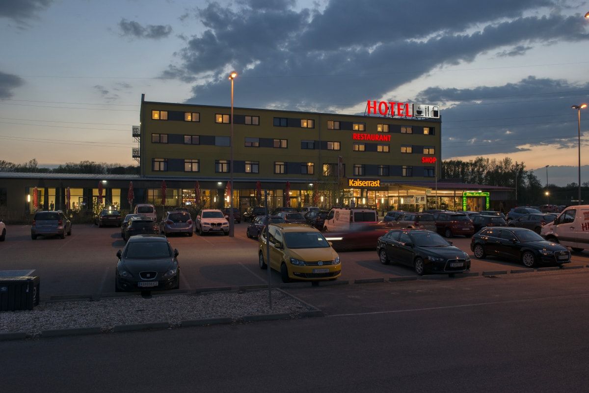 Večerní záběr hotelu s parkovištěm a auty před ním.