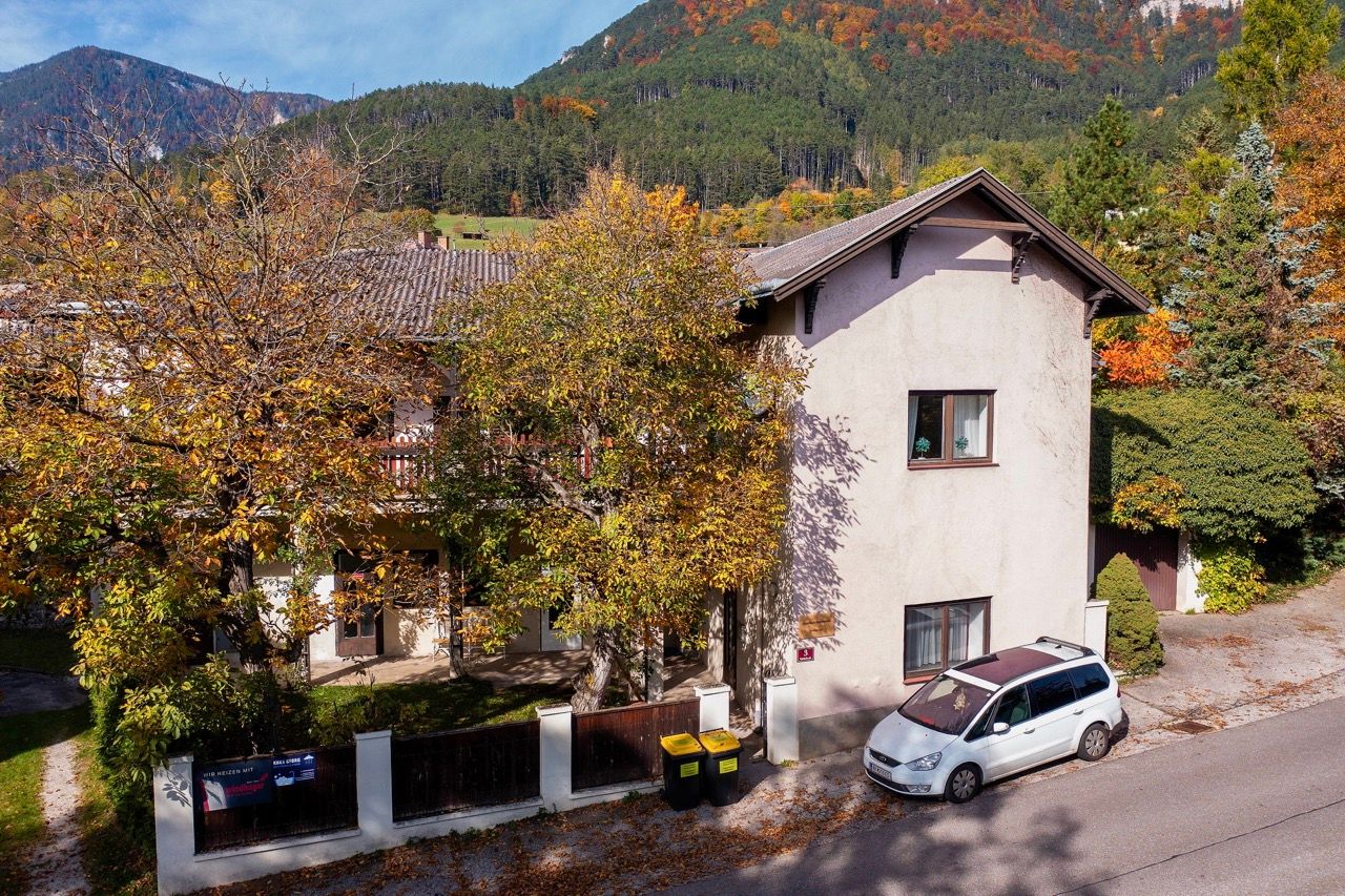 Dvoupatrová budova s podzimními stromy a zaparkovaným autem před ní, obklopená zalesněnými kopci.