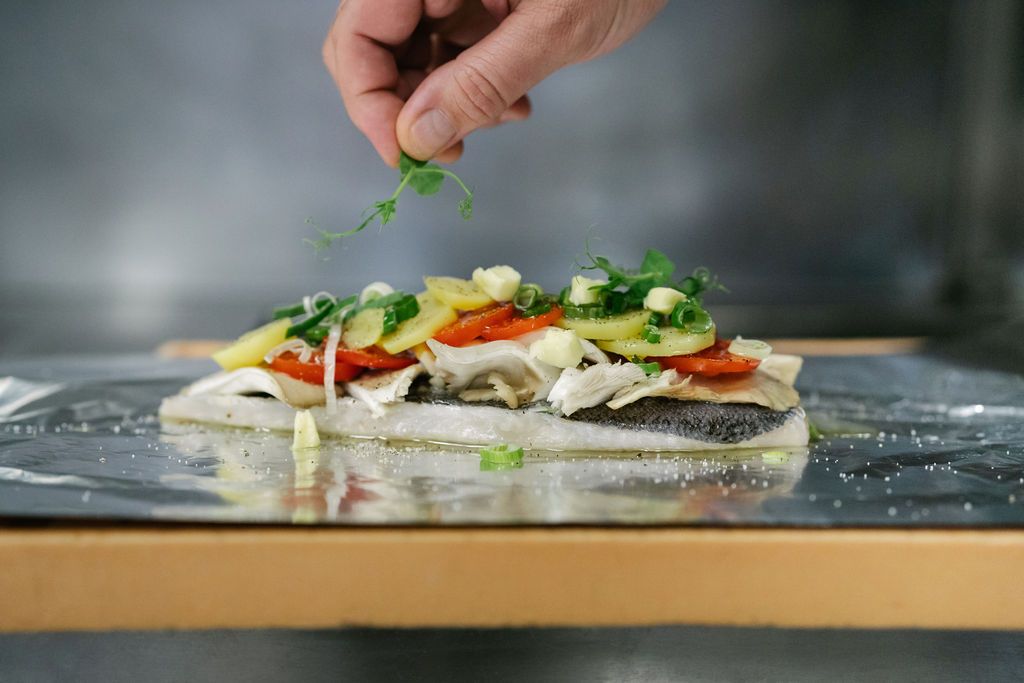 Rybí filet se zeleninou a bylinkami na alobalu, ruka přidává přílohu.