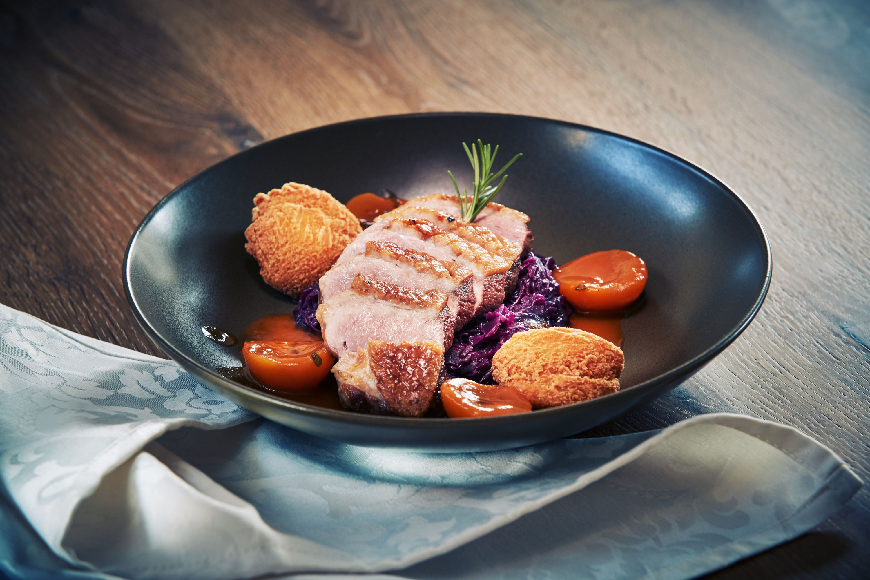 Gurmánský pokrm s kachními prsíčky, červeným zelím, bramborovými knedlíky a meruňkami na černém talíři.