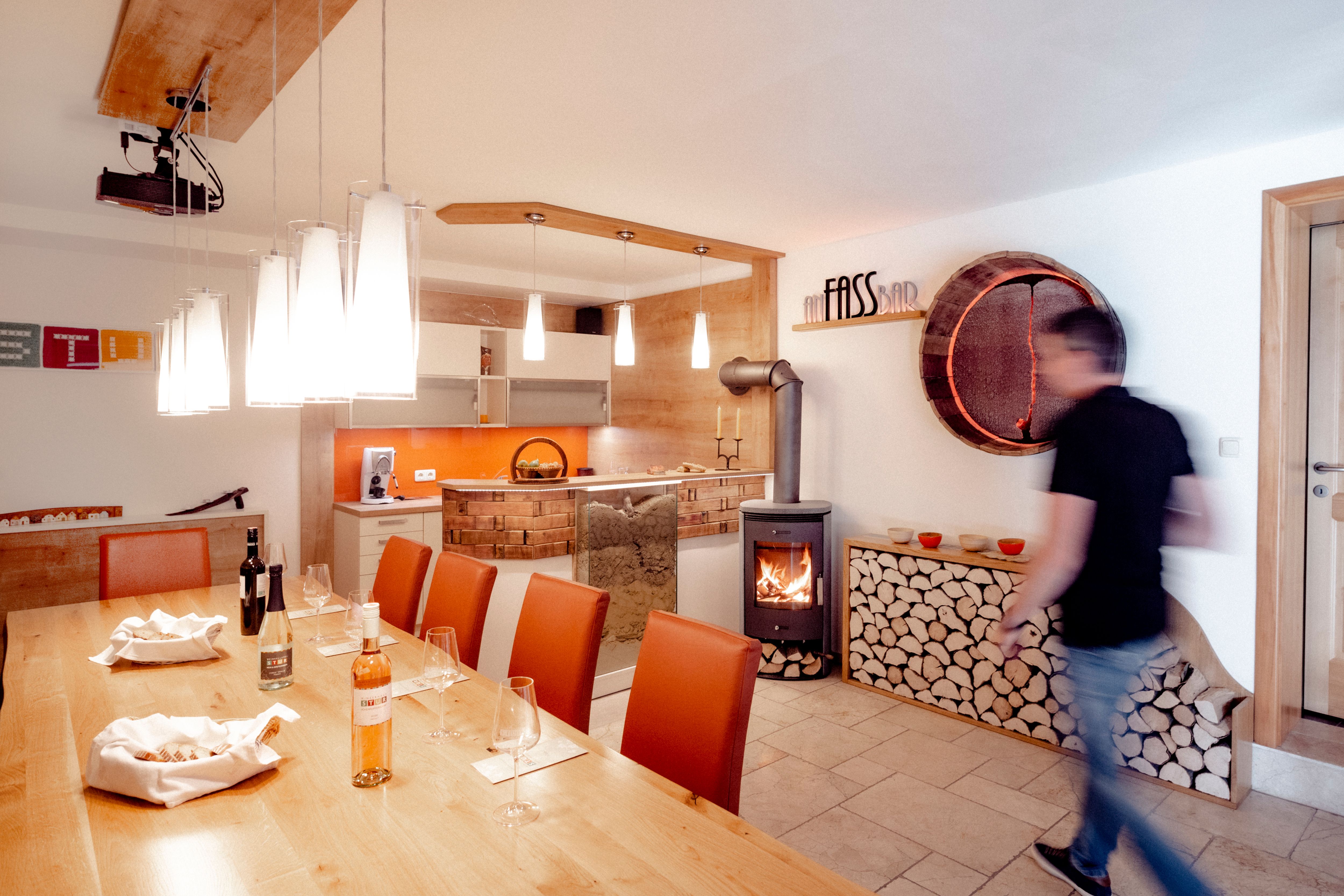 Degustační místnost s dřevěným stolem, sklenicemi na víno, krbem a moderním osvětlením.