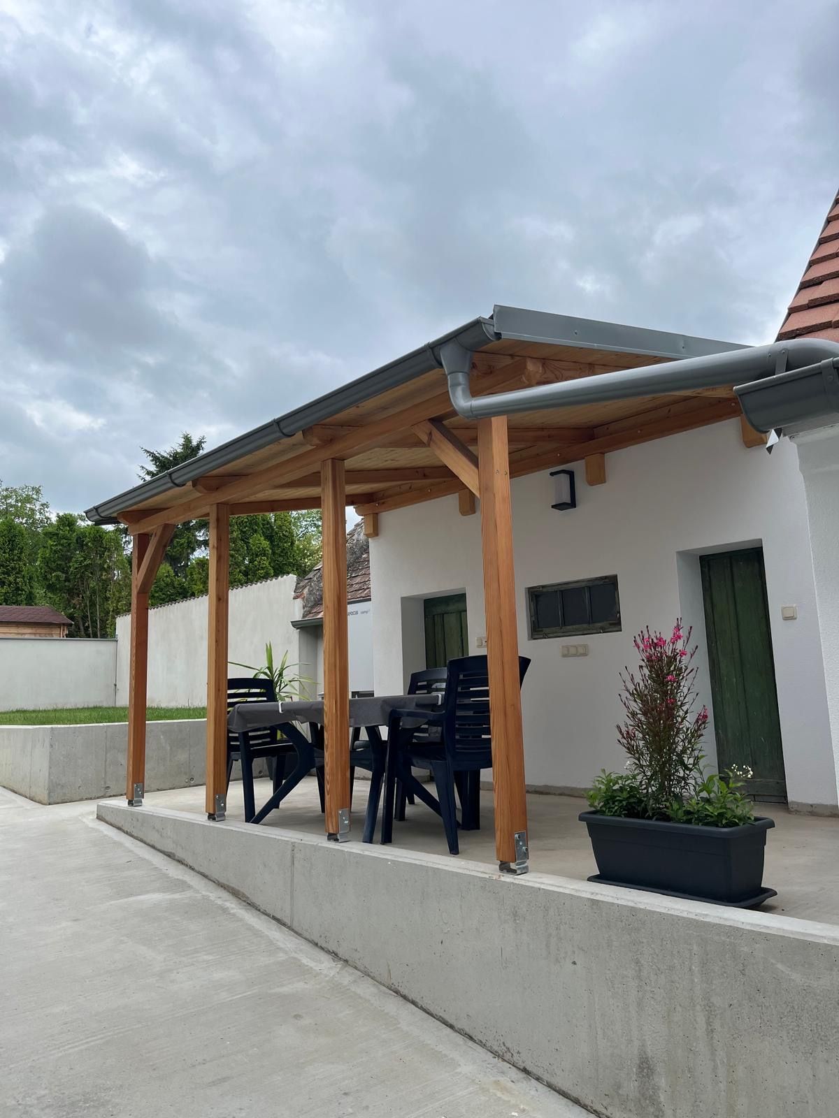 Krytá terasa se stolem a židlemi, obklopená rostlinami.