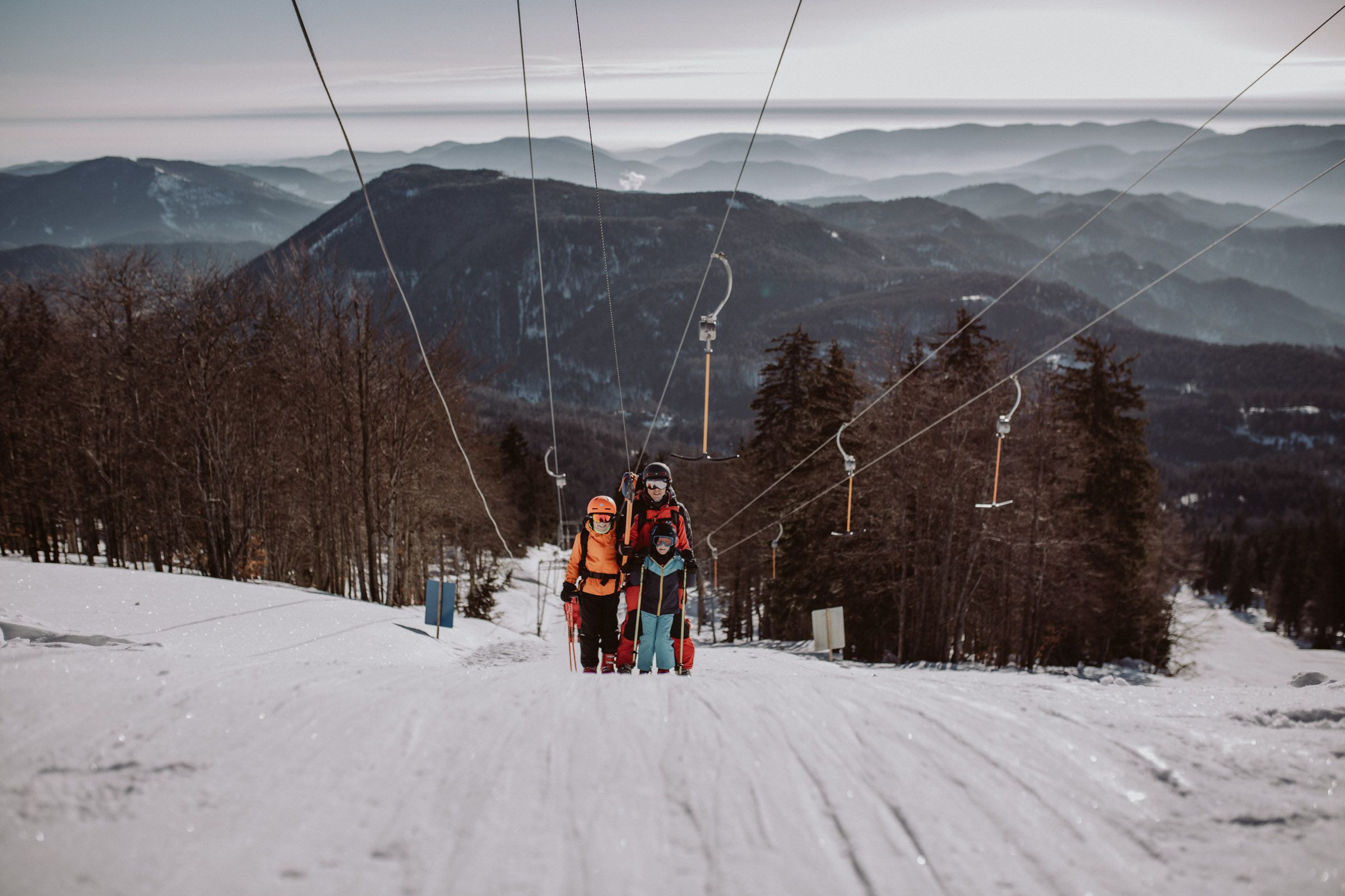 Tři lyžaři na vleku v lyžařské oblasti Unterberg, obklopeni zasněženými horami a stromy.