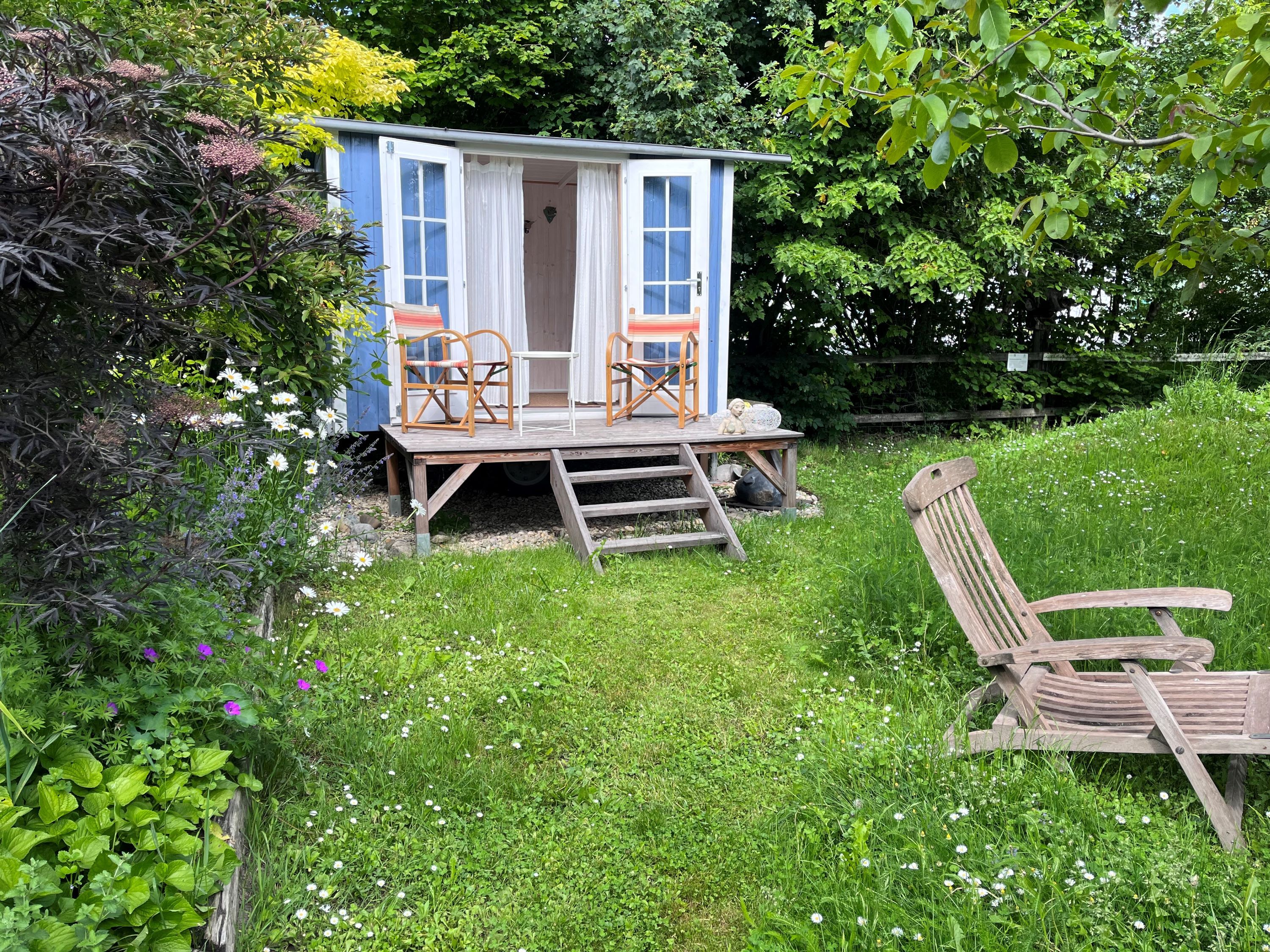 Modrý zahradní pavilon se dvěma židlemi na dřevěné verandě, obklopený zelenou loukou a barevnými květinami.