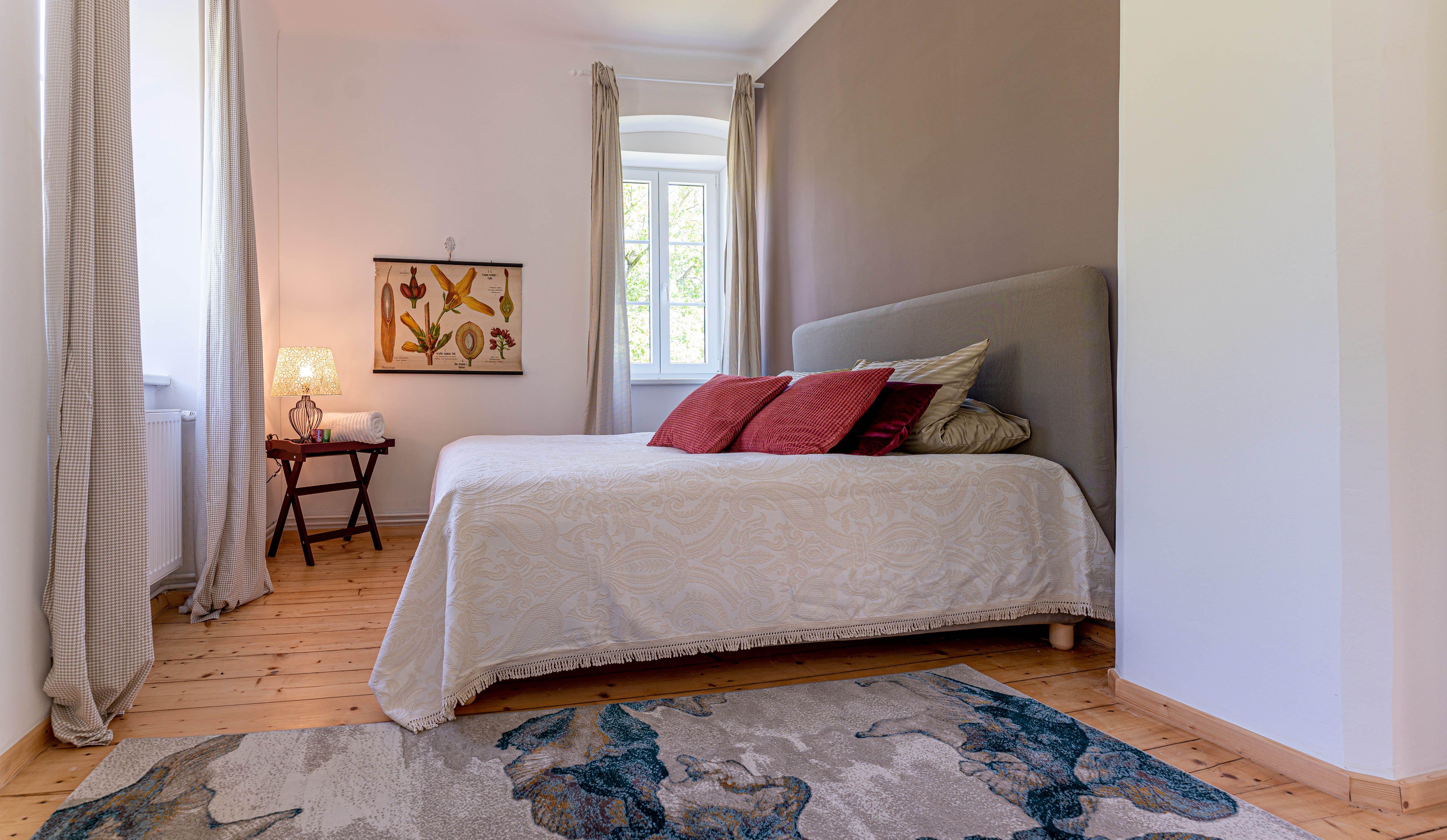 Ložnice s postelí, červenými polštáři, kobercem a oknem se závěsy.