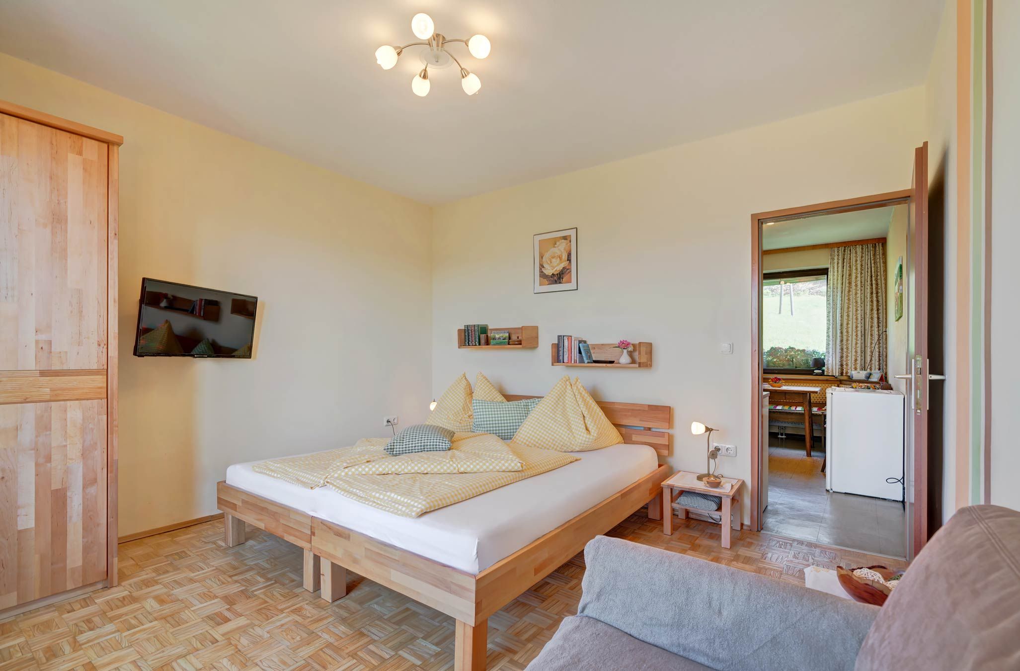 Útulná ložnice s dřevěnou podlahou, manželskou postelí se žlutým ložním prádlem, TV s plochou obrazovkou na stěně a otevřeným průchodem do kuchyně.