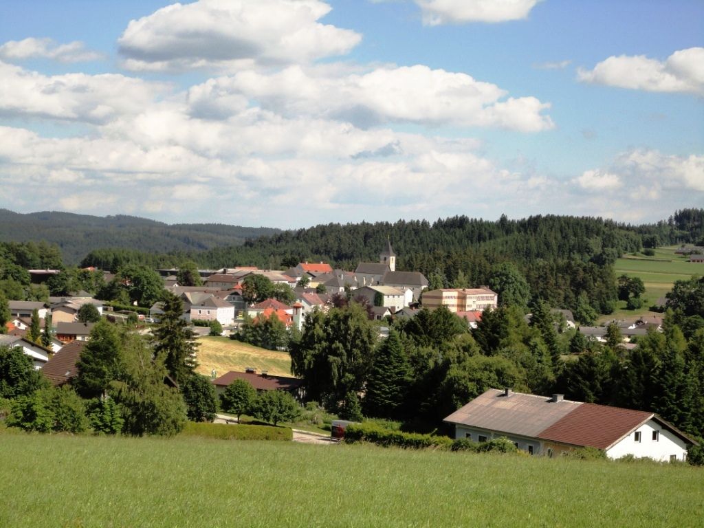 Panorama tržního městečka Martinsberg s kostelem a okolními domy, obklopeného lesy a poli pod modrou oblohou.