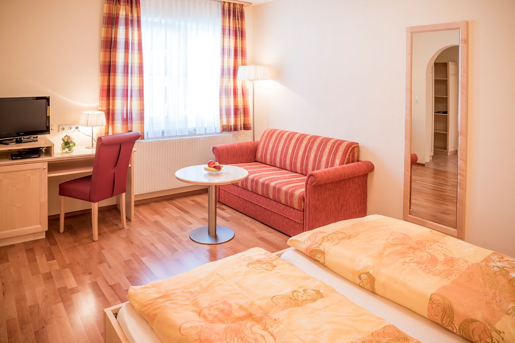 Útulný pokoj pro hosty s manželskou postelí, červenou pohovkou, psacím stolem, TV a velkým zrcadlem.