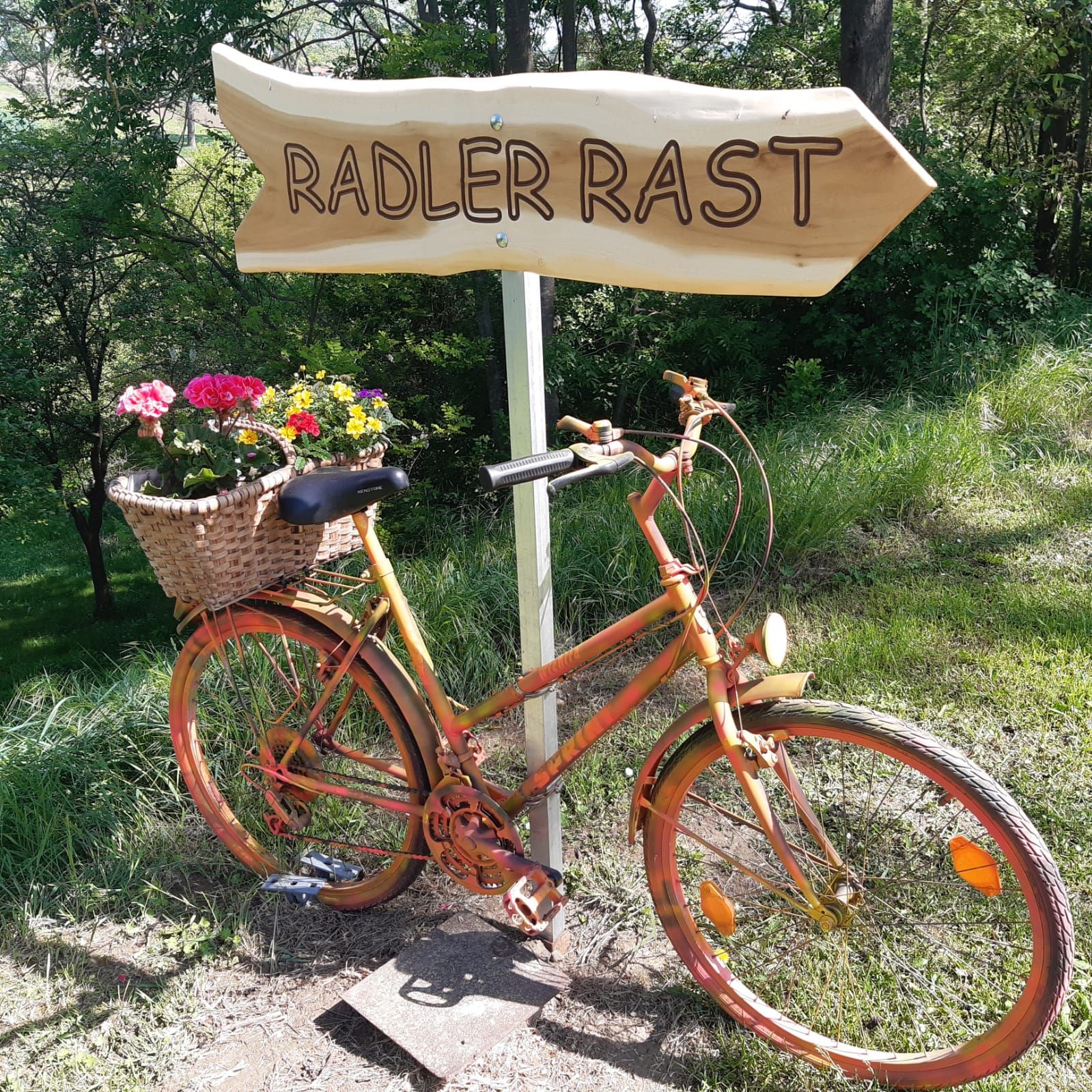 V zeleném prostředí stojí oranžové kolo s košíkem na květiny vedle dřevěné cedule s nápisem „Radler Rast“.